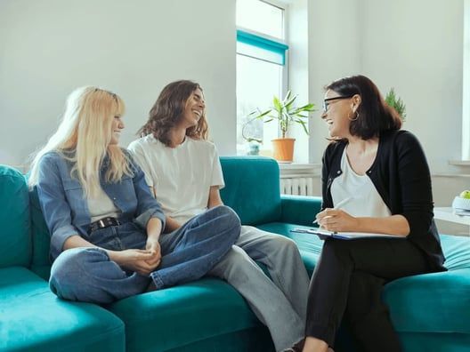 A couple sits with a therapist on a teal couch in a sunlit room. The therapist, holding a clipboard, smiles while talking.