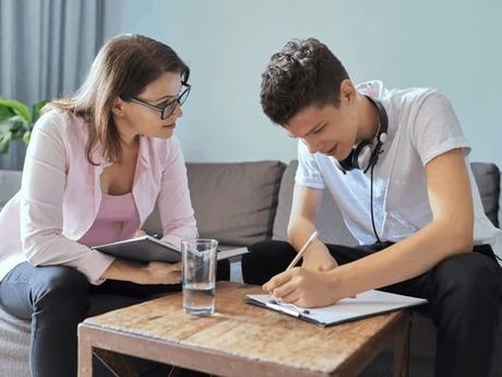 A woman in glasses and a pink blazer sits with a teenage boy writing on a clipboard. They are indoors, possibly during a therapy session.