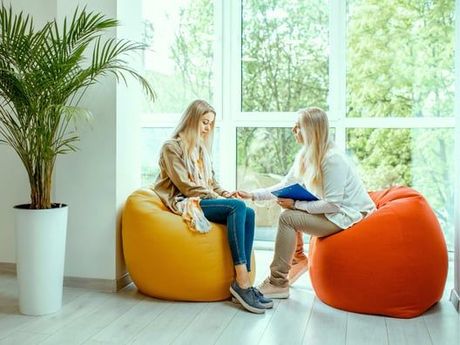 Woman in therapy session, sitting on a yellow beanbag, holding hands with a therapist on an orange beanbag in front of a window.