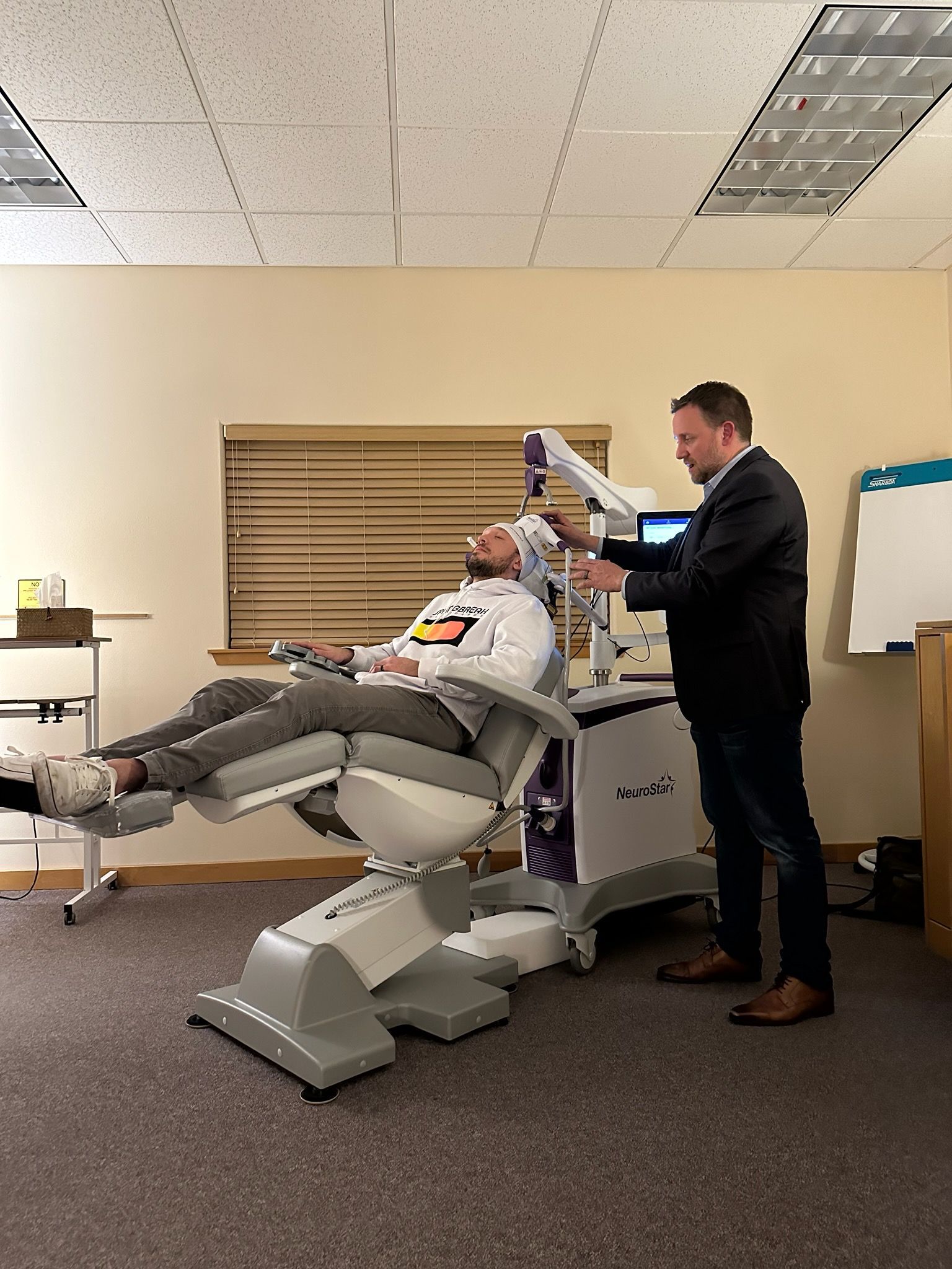 A woman sits in a medical chair, smiling at a man holding a tablet. Purple and white medical equipment is in a bright, modern room.
