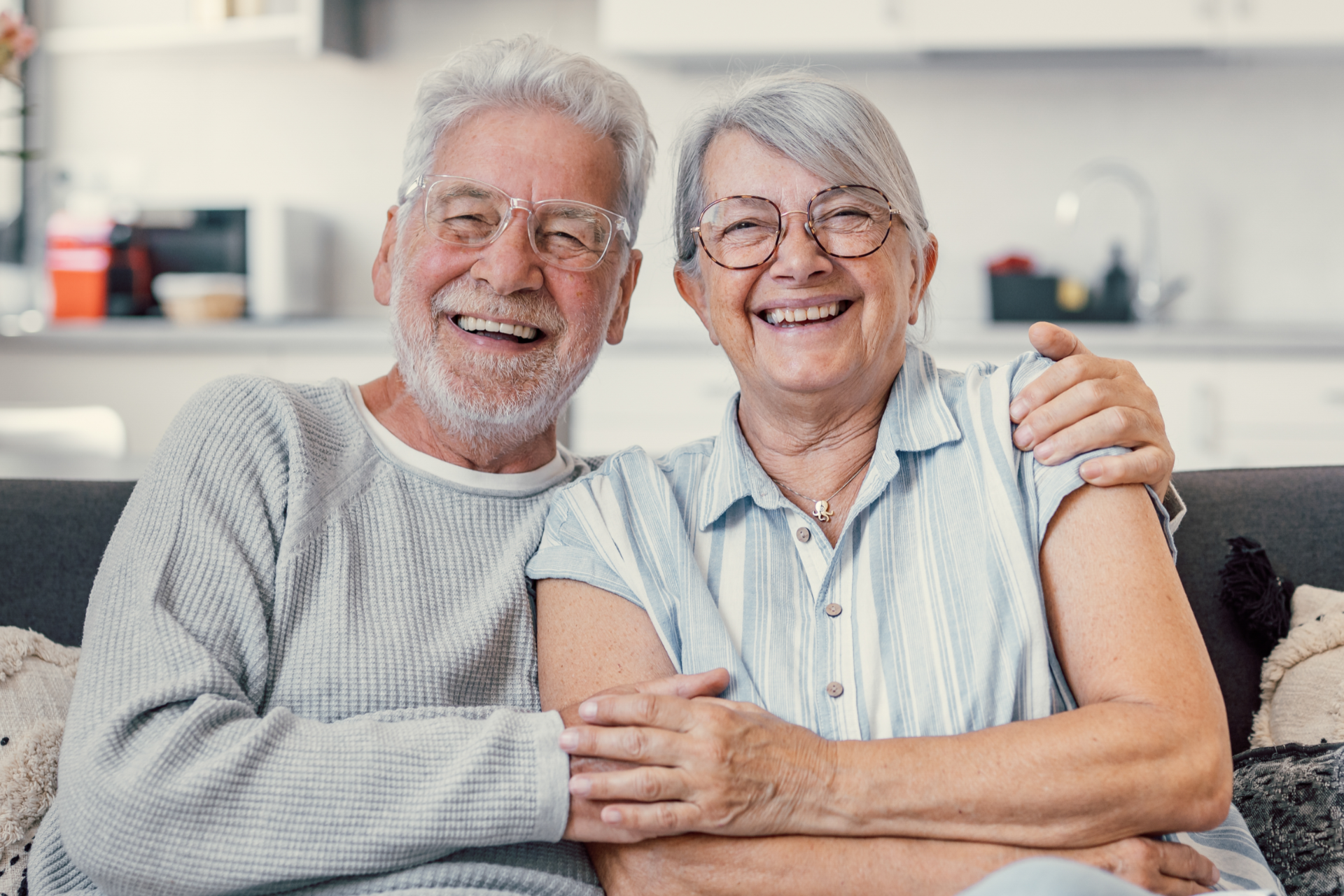 Smiling older couple sitting on a couch, embracing in a bright living room.