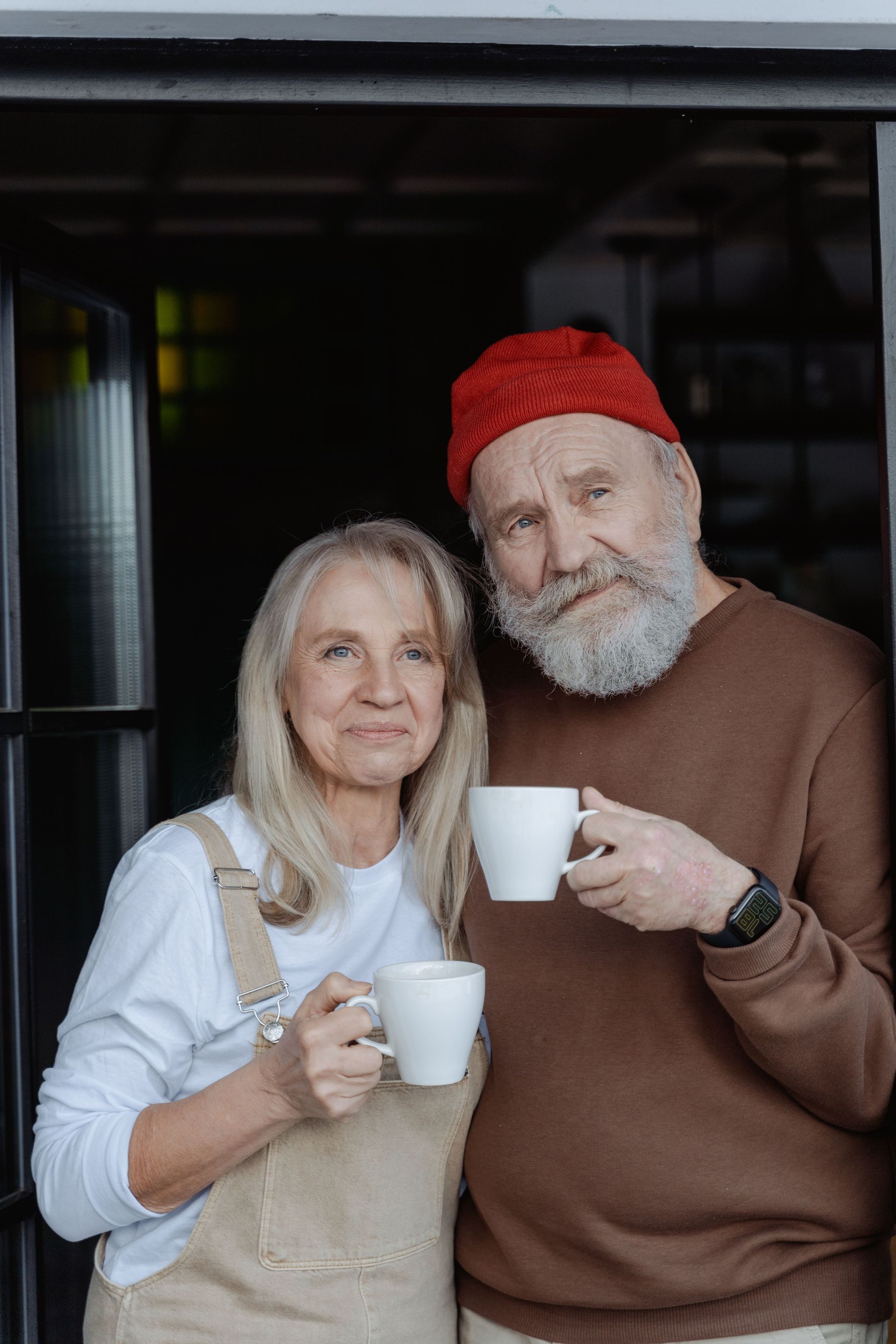 Two people standing together indoors, each holding a white mug, one in a red hat and the other in a cream sweater