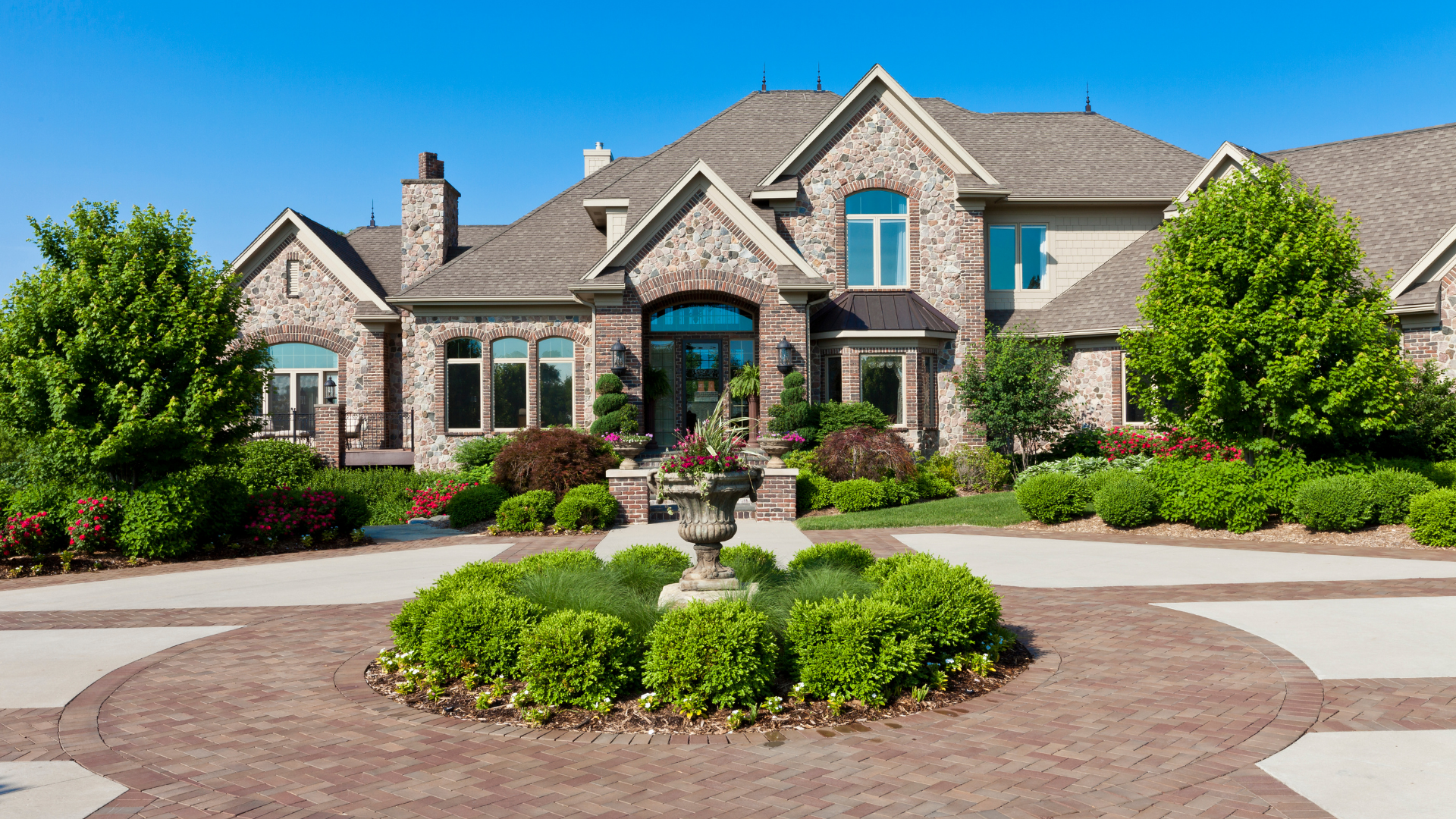 Large stone suburban house with landscaped front yard and circular driveway under a clear blue sky