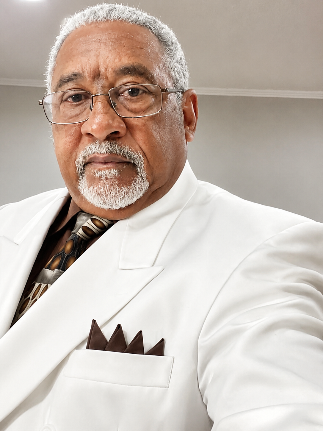 Portrait of a man in a white suit and glasses, looking at the camera against a gray background