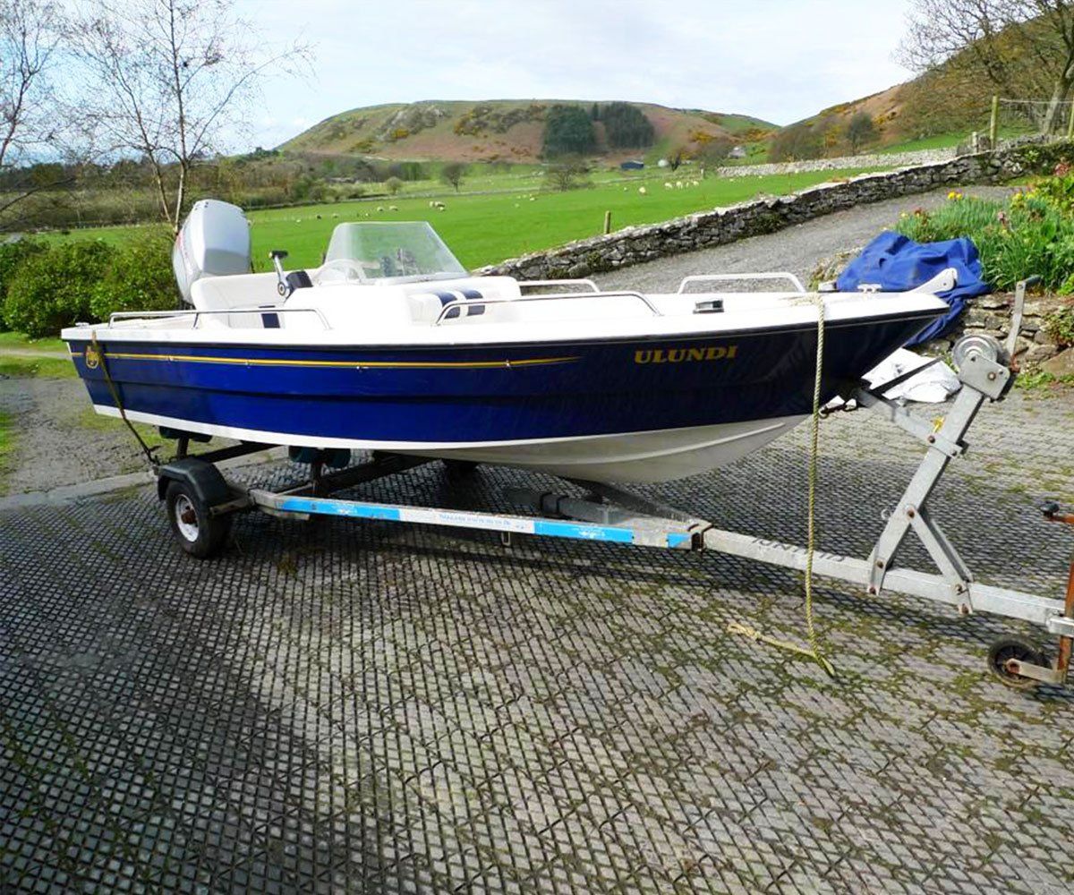 Blue and white motorboat on a trailer, parked on a gravel driveway, with a green hill in the background.