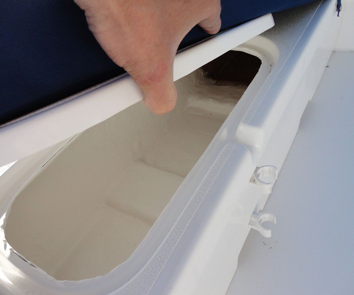 Hand lifting a white lid to reveal an open, rectangular storage compartment in a boat.