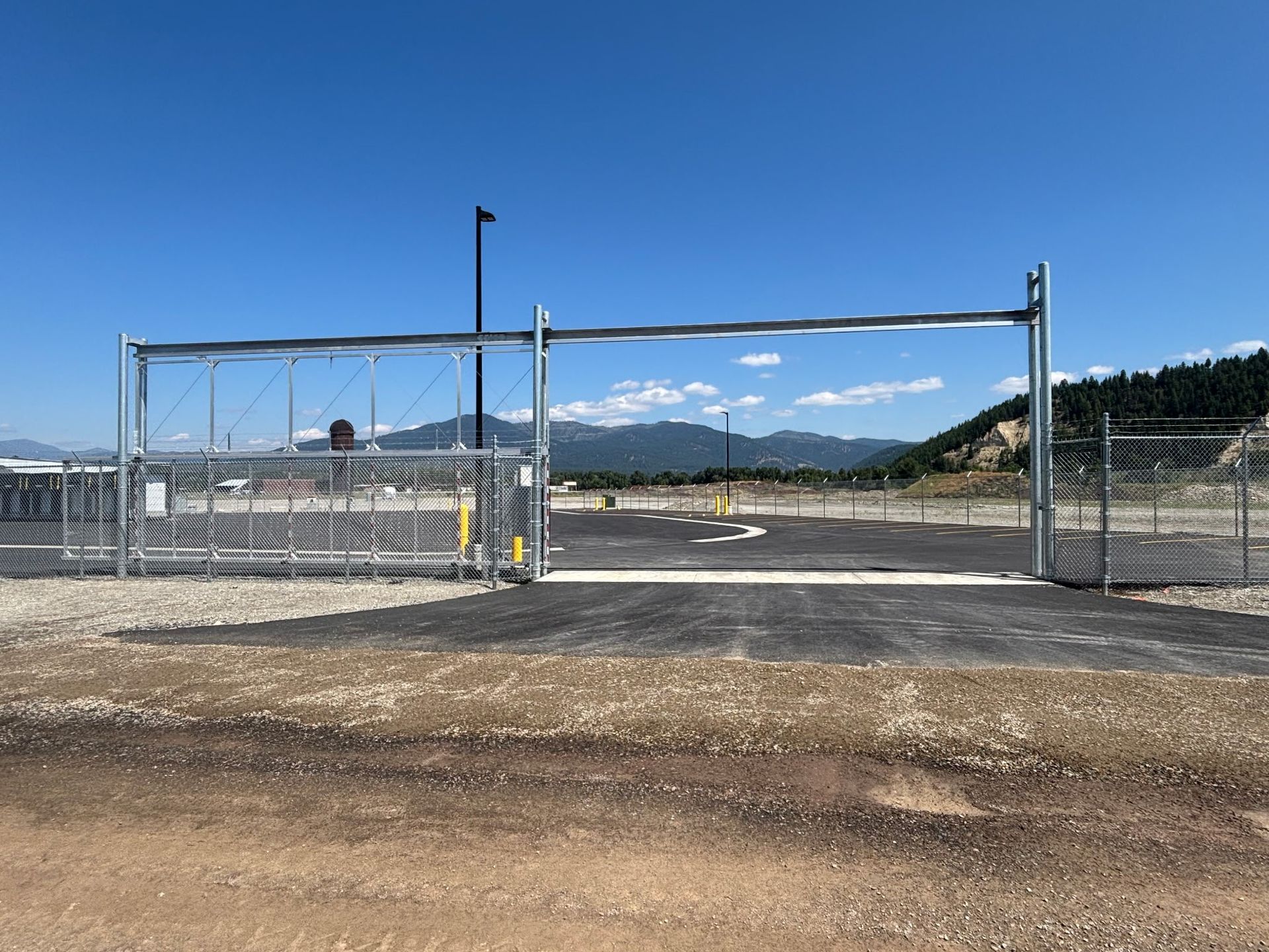 Open metal gate on a paved road, leading to a parking area. Mountains and blue sky in the background.