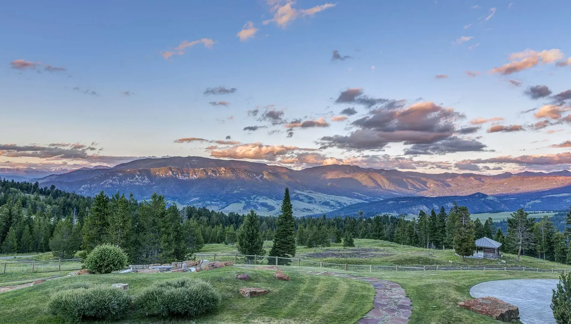 Rolling green landscape with mountains and trees under a cloudy sunset sky.