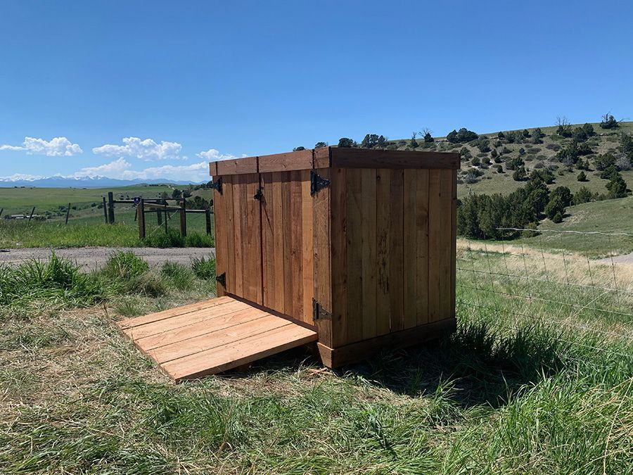 A wooden shed with a ramp is sitting in the middle of a grassy field.
