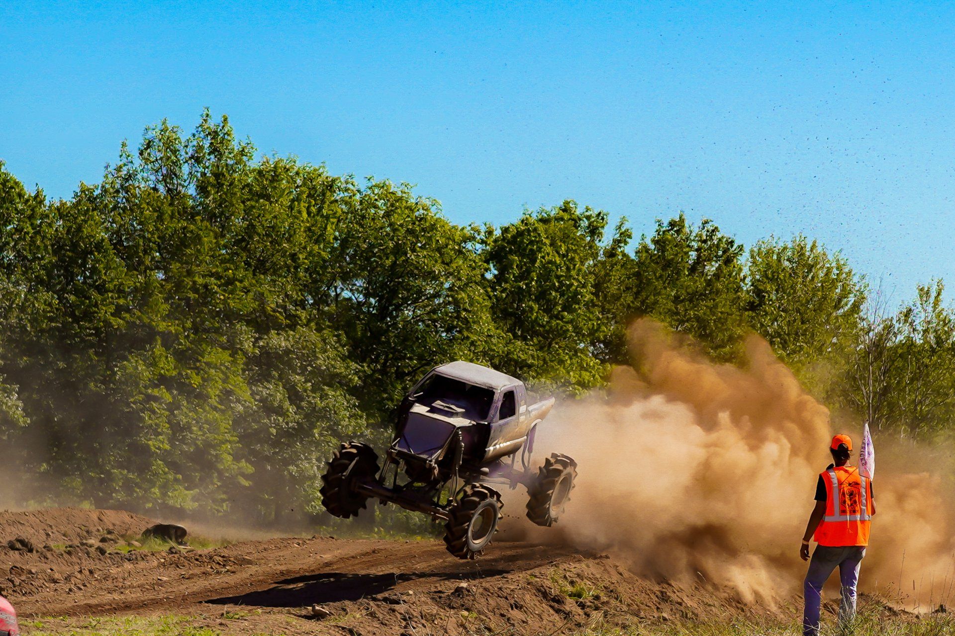 Off-road buggy kicking up dust on a dirt track as a person in an orange vest watches nearby.