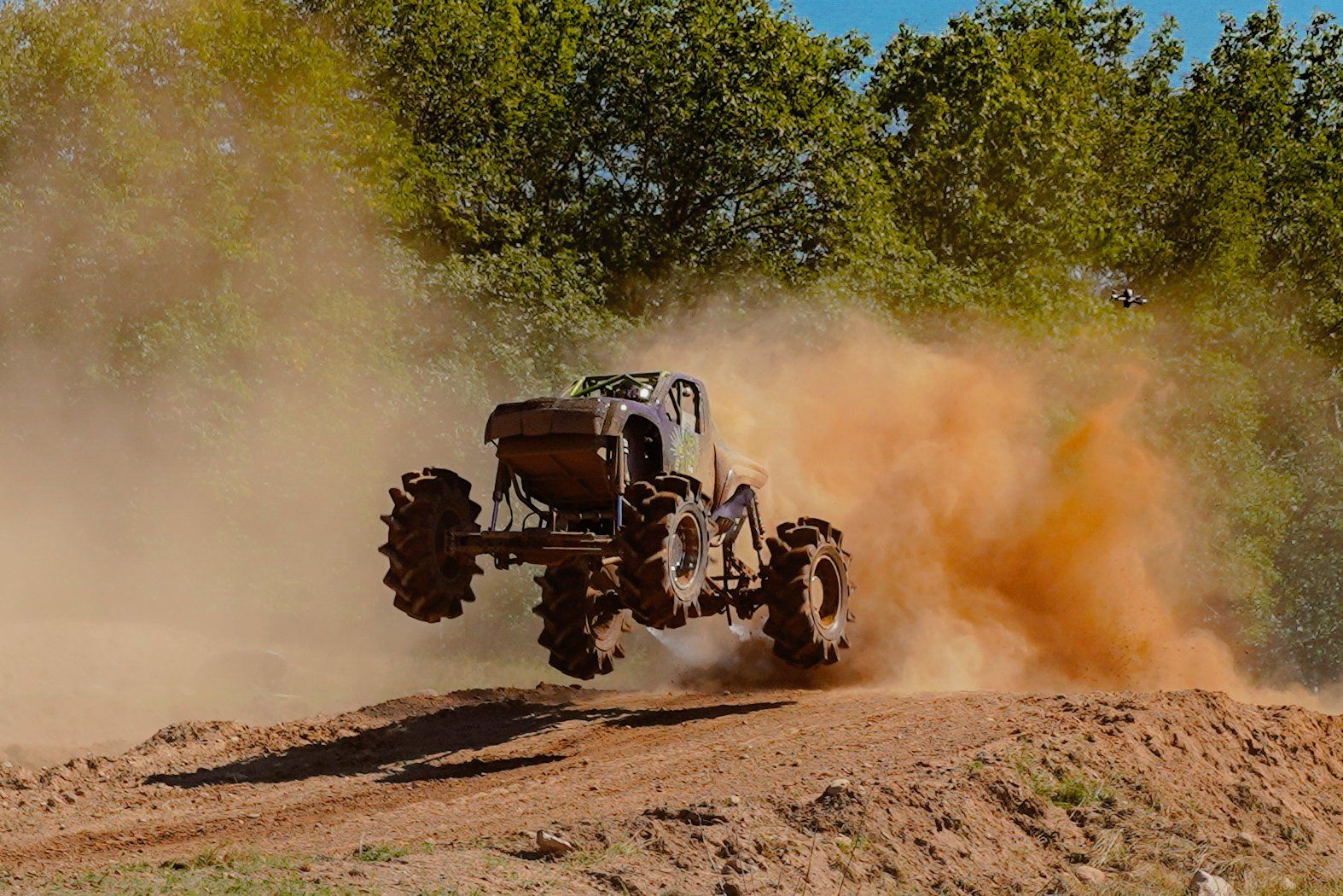 Mud-covered off-road truck airborne on a dirt track, kicking up dust near trees