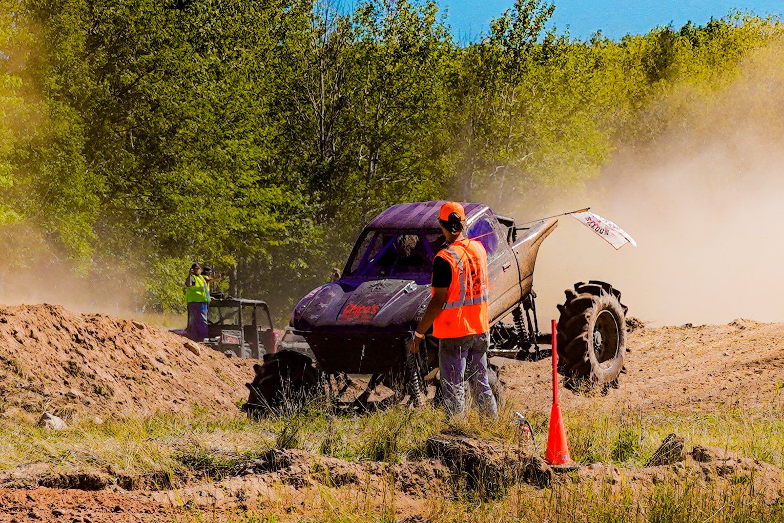 Off-road buggy stopped in a dusty field, with two workers beside it and trees in the background.