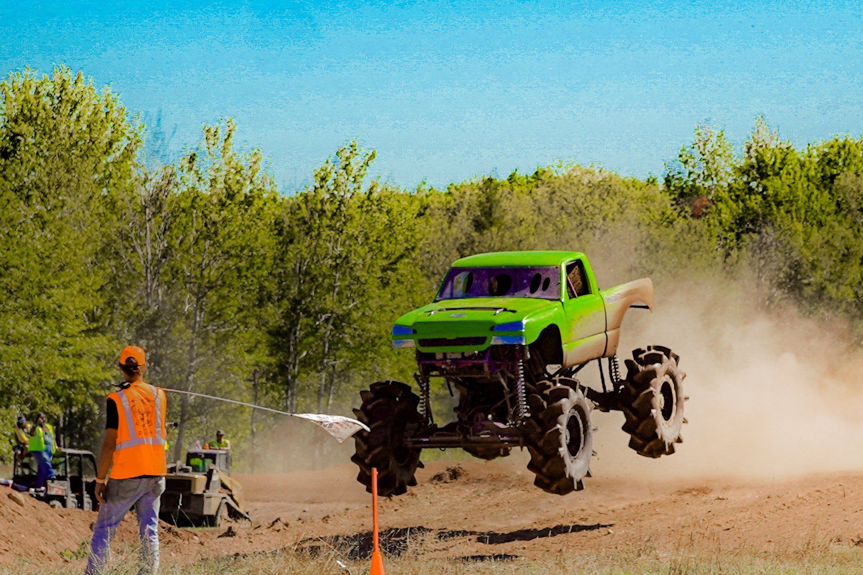 Bright green monster truck airborne on a dirt track, kicking up dust, with a marshal in orange nearby
