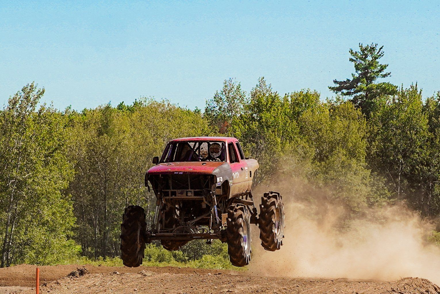 Red off-road truck airborne on a dirt trail, kicking up dust with trees in the background