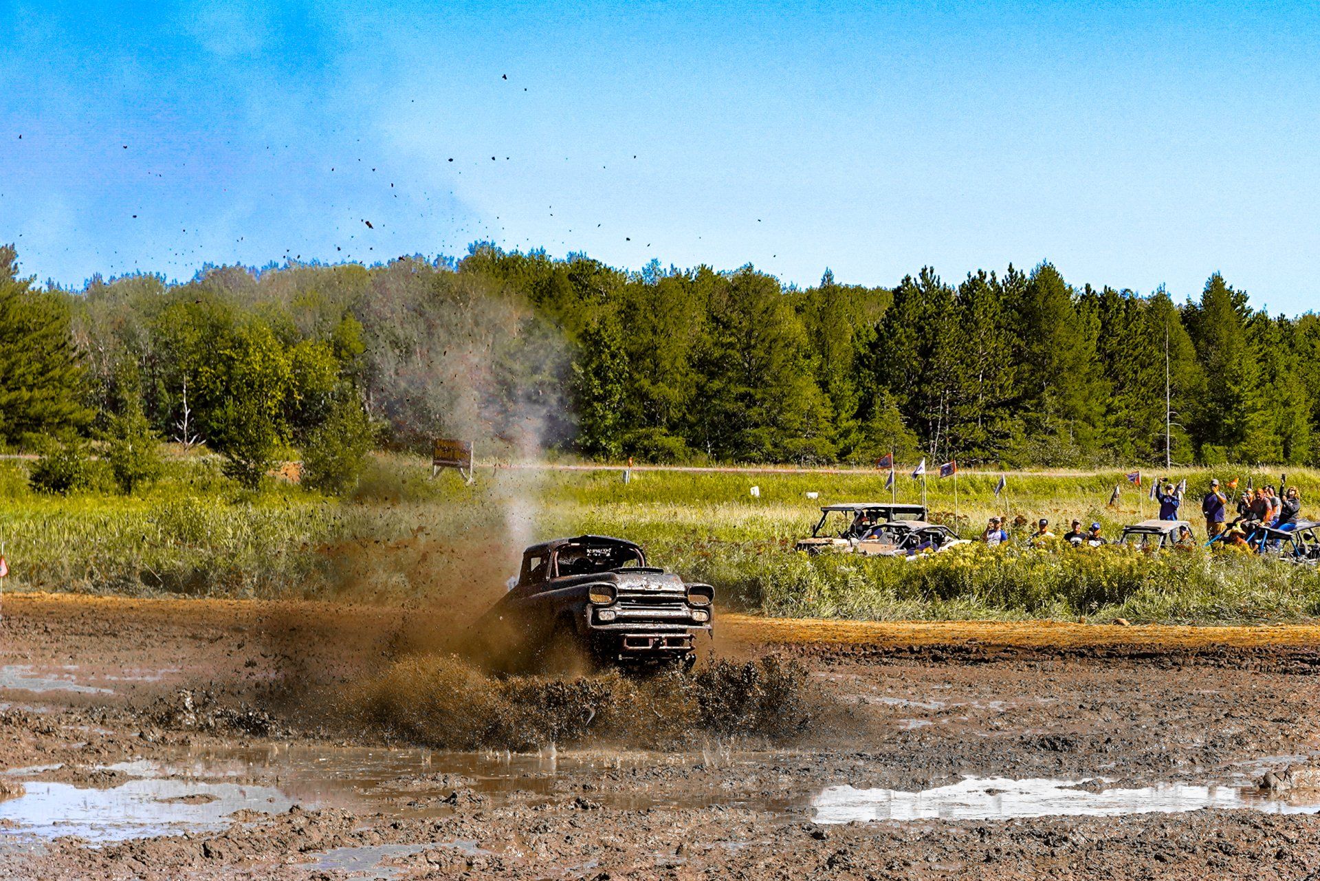 Off-road truck splashing through muddy water at a dirt track, with spectators and trees in the background