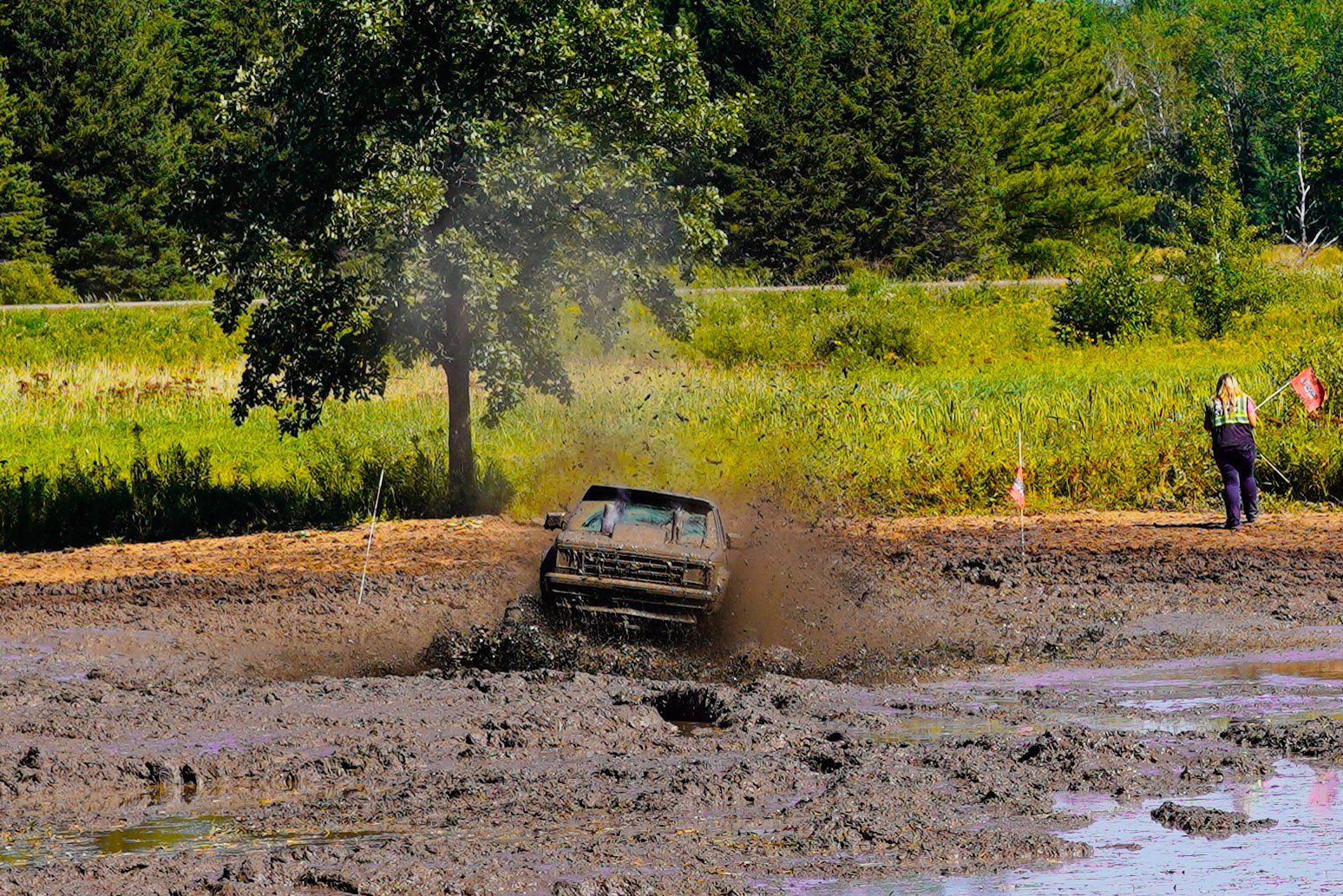 Off-road truck splashing through muddy water in a wooded field, with two spectators nearby