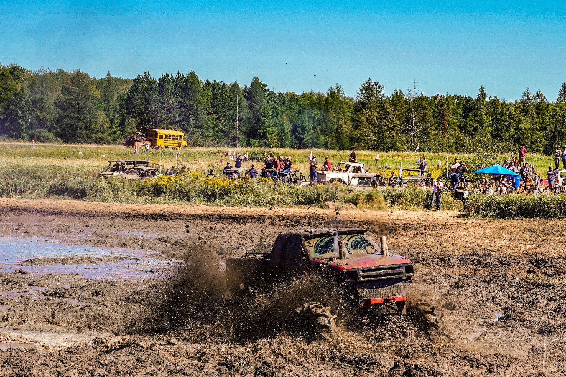 Off-road truck splashing through a muddy field at a rally, with spectators and trees in the background