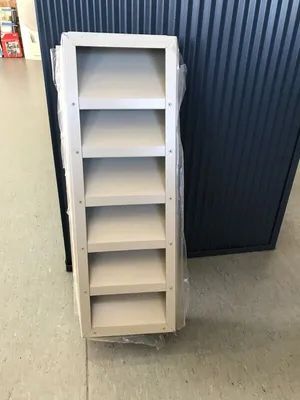 A narrow, beige wooden bookshelf with six shelves, wrapped in plastic, against a corrugated blue wall. — R&M Sheetmetal Nowra Metal Roofing in South Nowra, NSW