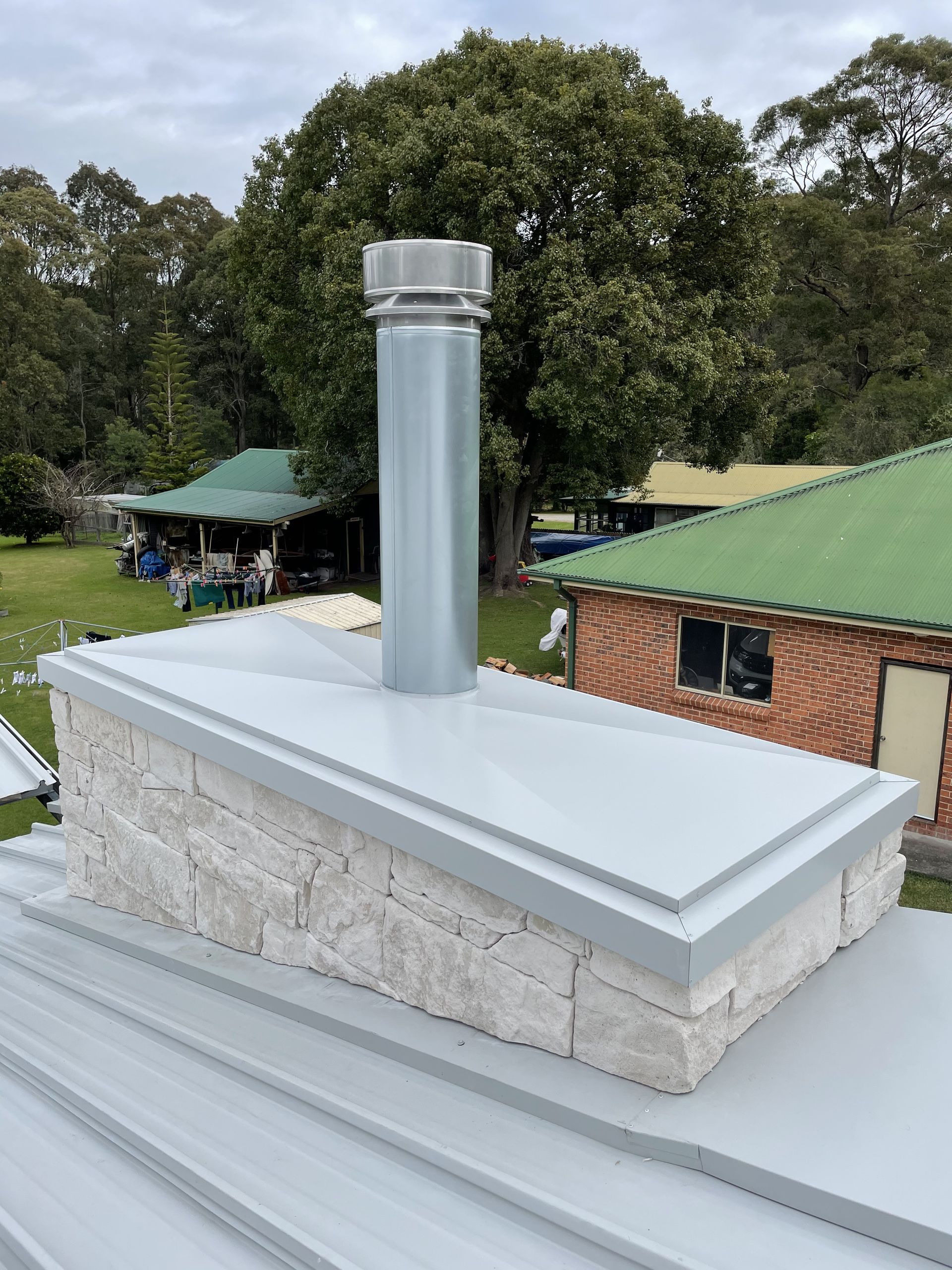 Chimney on a grey roof, with a metal flue. A brick house and trees are in the background. — R&M Sheetmetal Nowra Metal Roofing in South Nowra, NSW