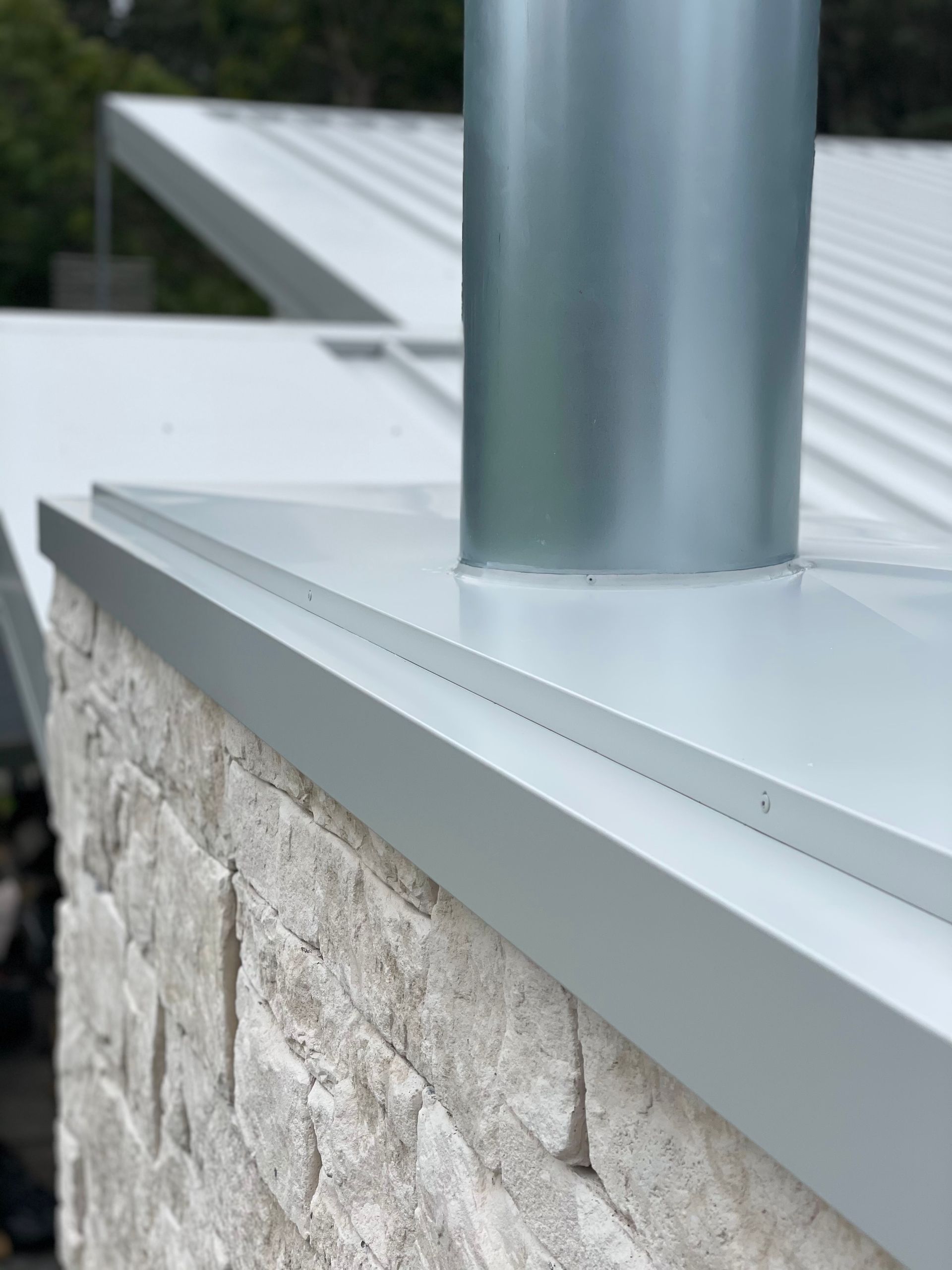 Metal chimney rising from a white flat roof against a stone brick wall. — R&M Sheetmetal Nowra Metal Roofing in South Nowra, NSW