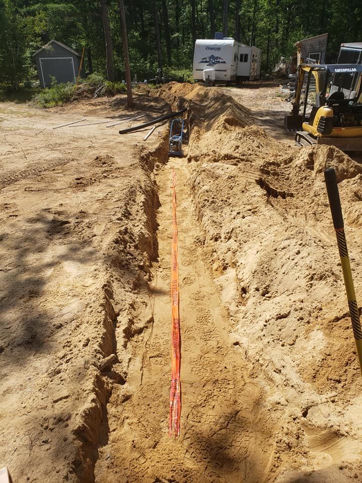 Trench in sandy soil with orange utility line; excavator and trailer in background.