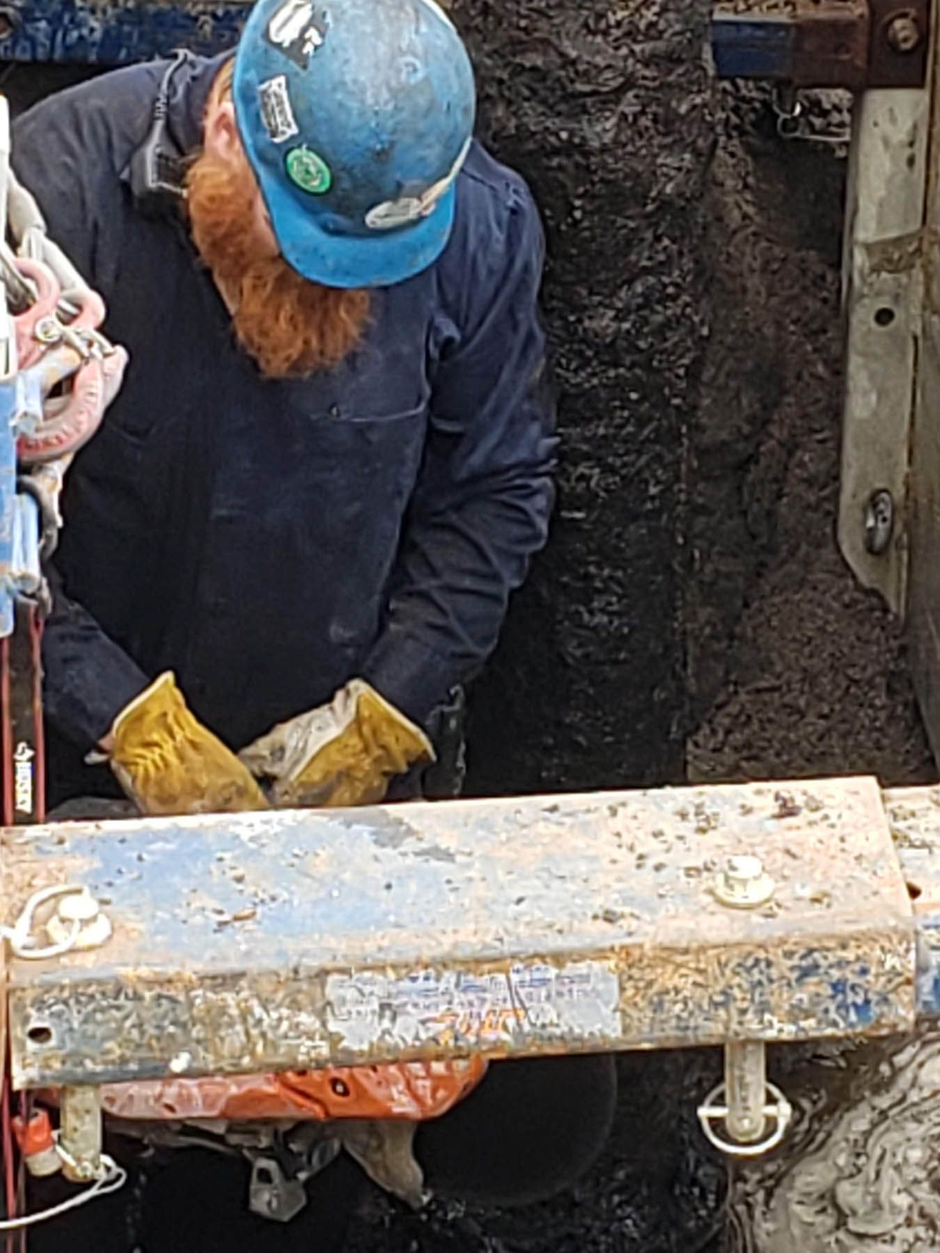 Man in blue hat and work clothes inspecting a dark hole, dirty setting.