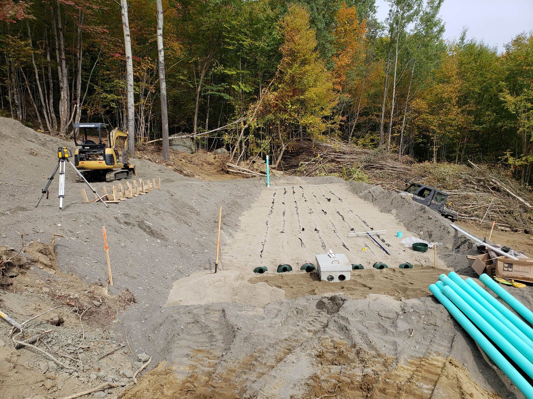 Construction site: excavation for septic system installation, pipes, gravel, mini excavator, forested background.
