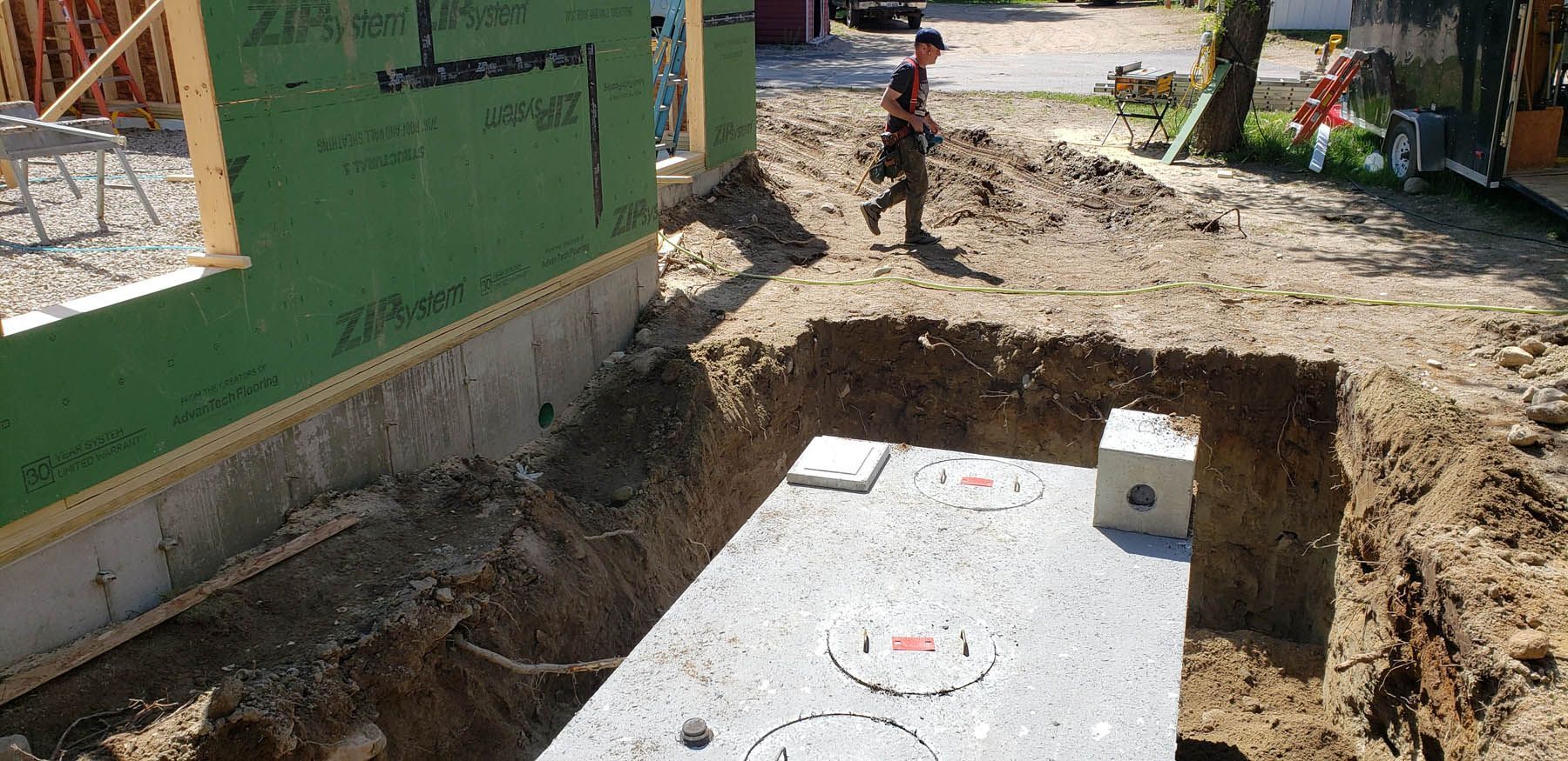 Construction site with a man walking near a buried septic tank. The foundation of a house is also visible.