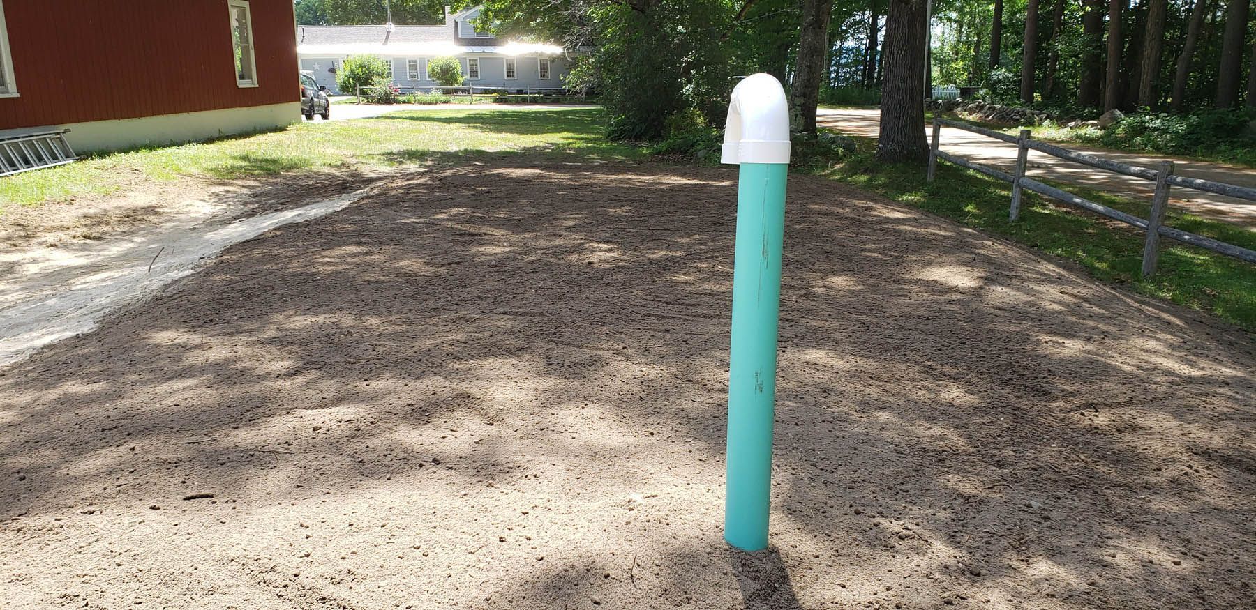 A green pipe with a white cap in a gravel area next to a path, buildings, and trees.