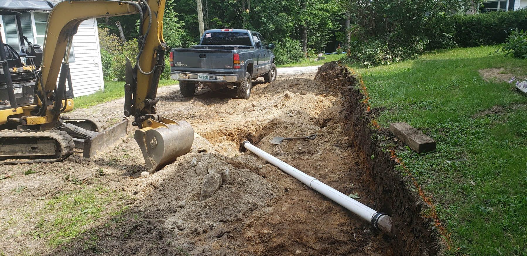 An excavator digging a trench, a white pipe in the trench, and a truck on a gravel driveway.