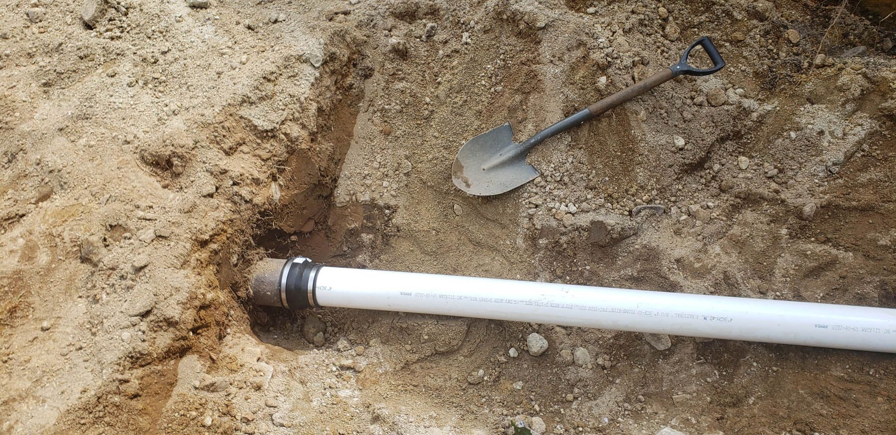 A shovel next to a white PVC pipe buried in dirt.
