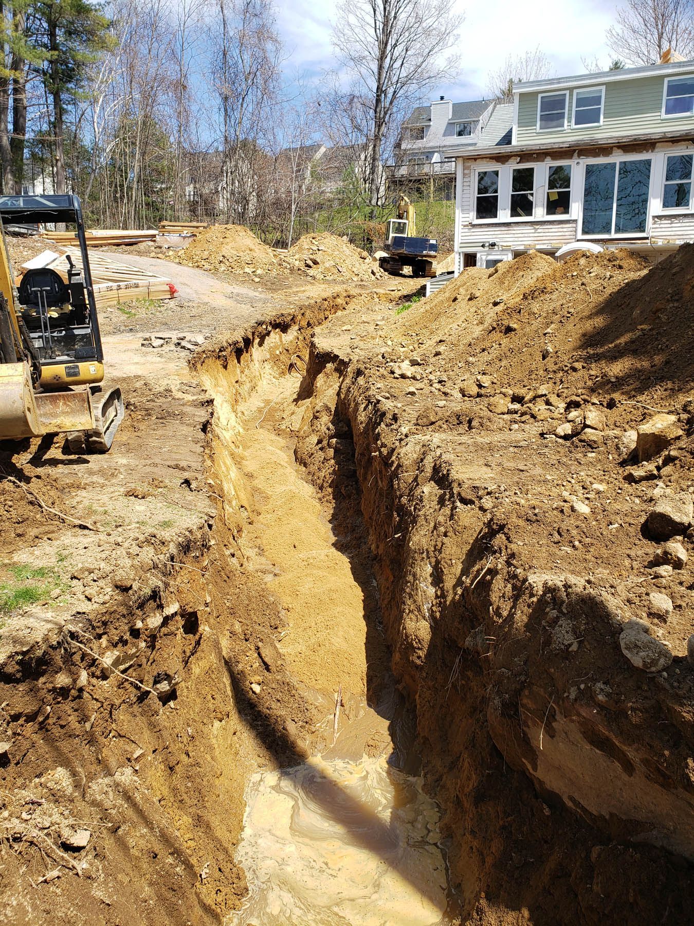 Excavation site with a long trench. A backhoe is parked on the left. A house is in the background.