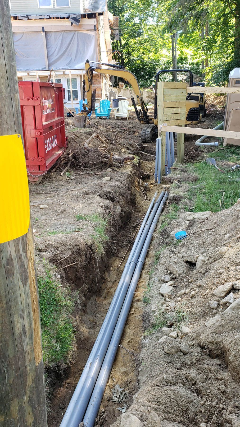 Construction site with a trench, PVC pipes, and an excavator, near a building.