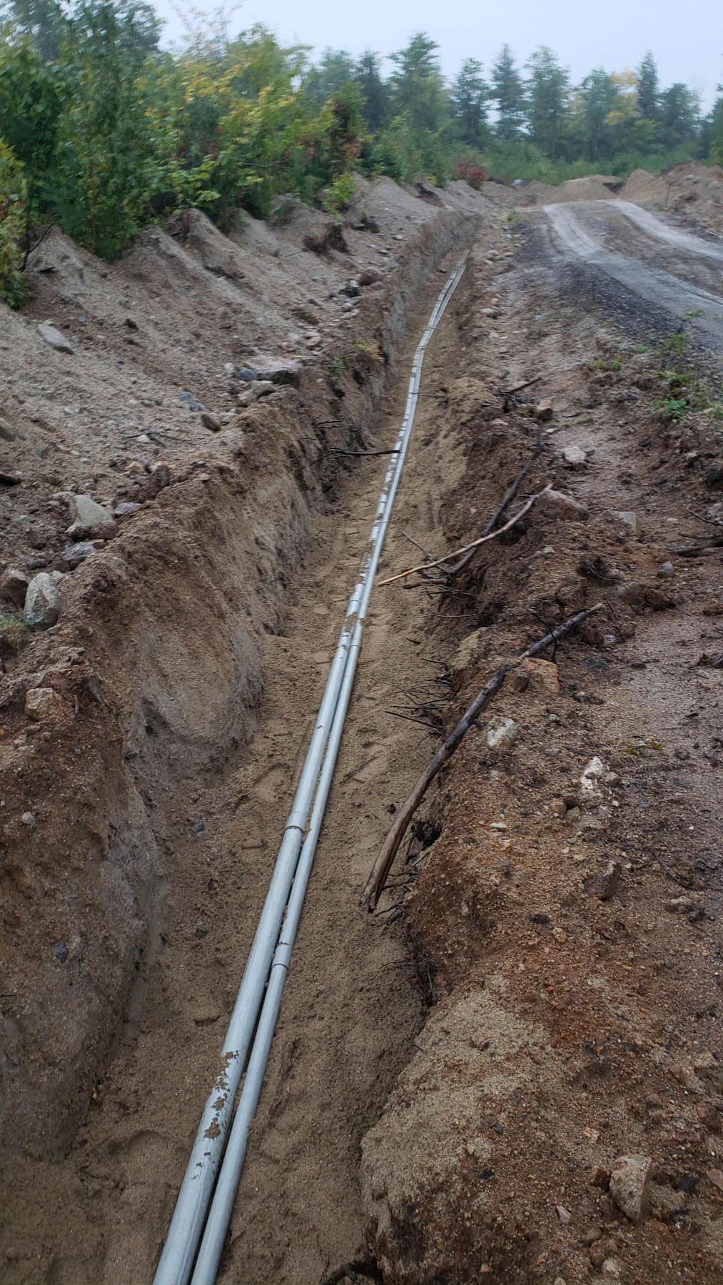 A narrow trench dug into dirt, holding two grey pipes; a hillside with trees is in the background.