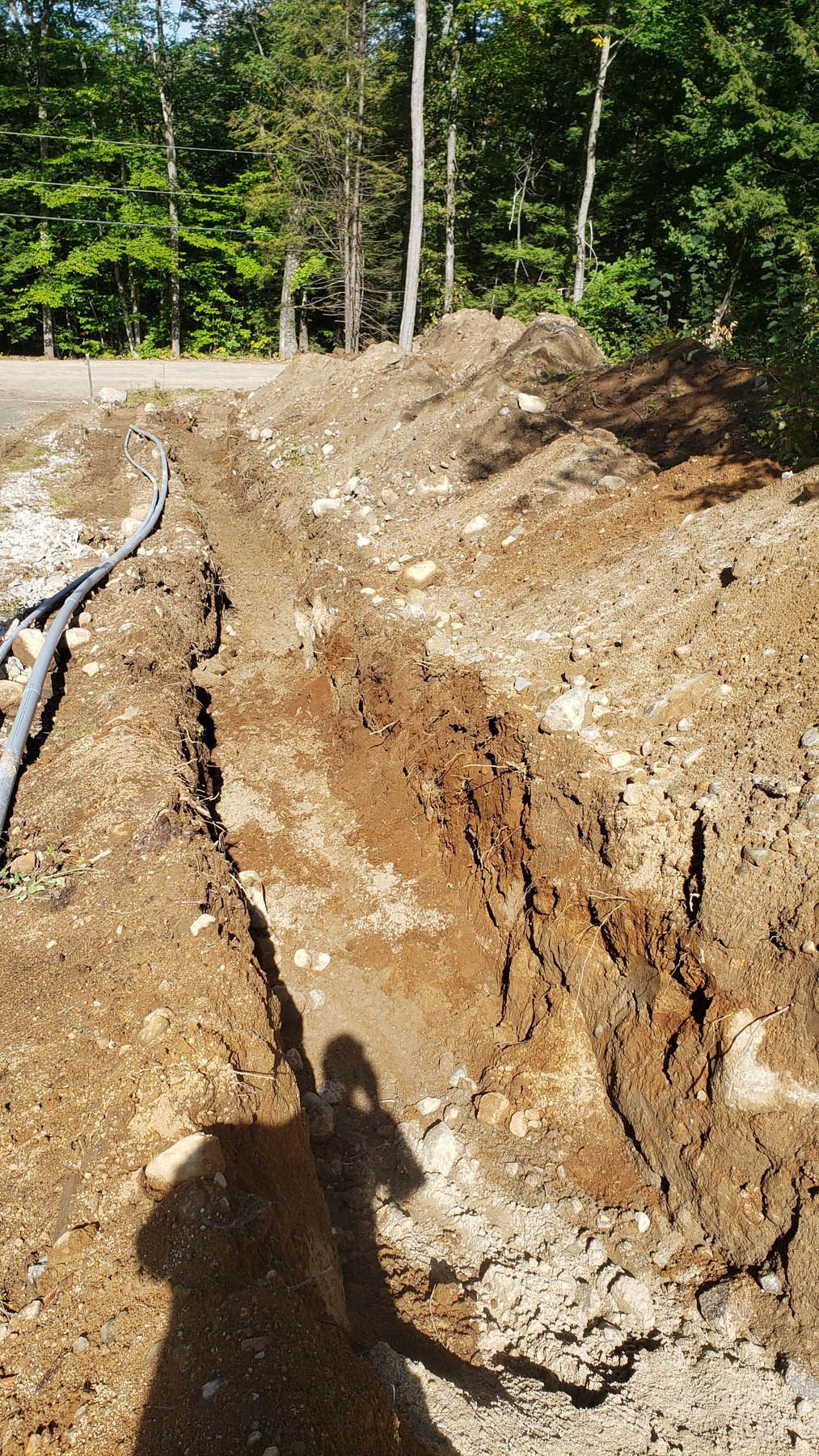 A trench dug in dirt, possibly for utility lines, with a pile of dirt in a wooded area.
