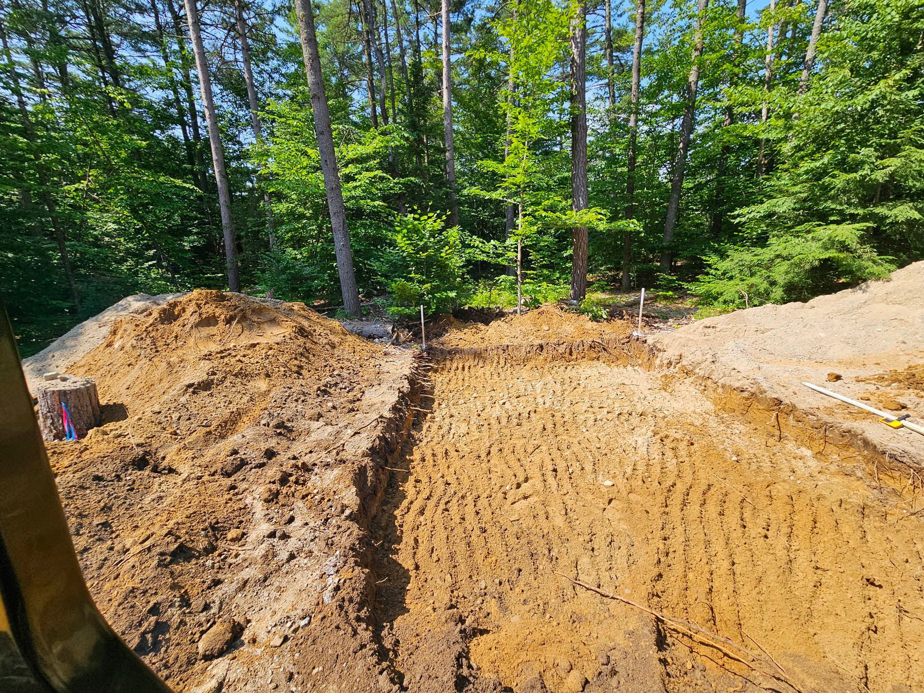 Excavated ground in a wooded area, showing dug trenches and a pile of dirt, ready for construction.