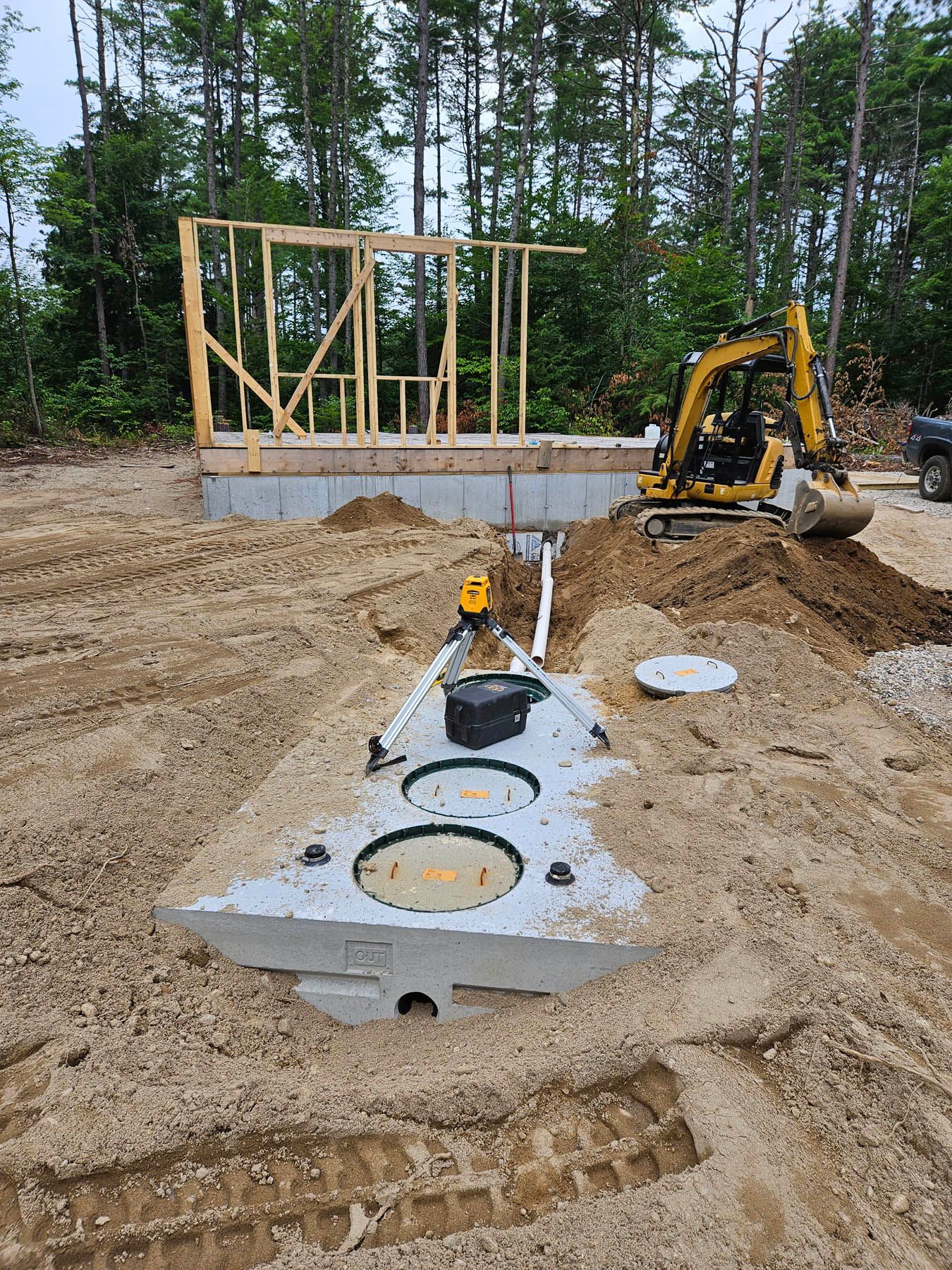 Construction site: septic system installation near a building frame; a yellow excavator is present.