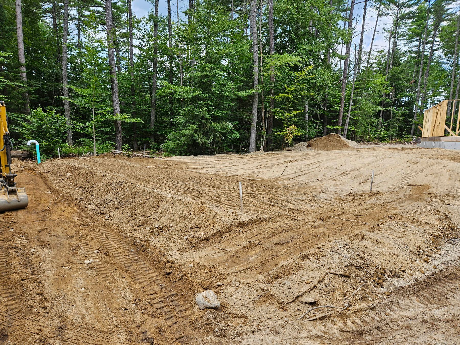 Graded dirt construction site, with tire tracks and utility pipe, trees in the background.