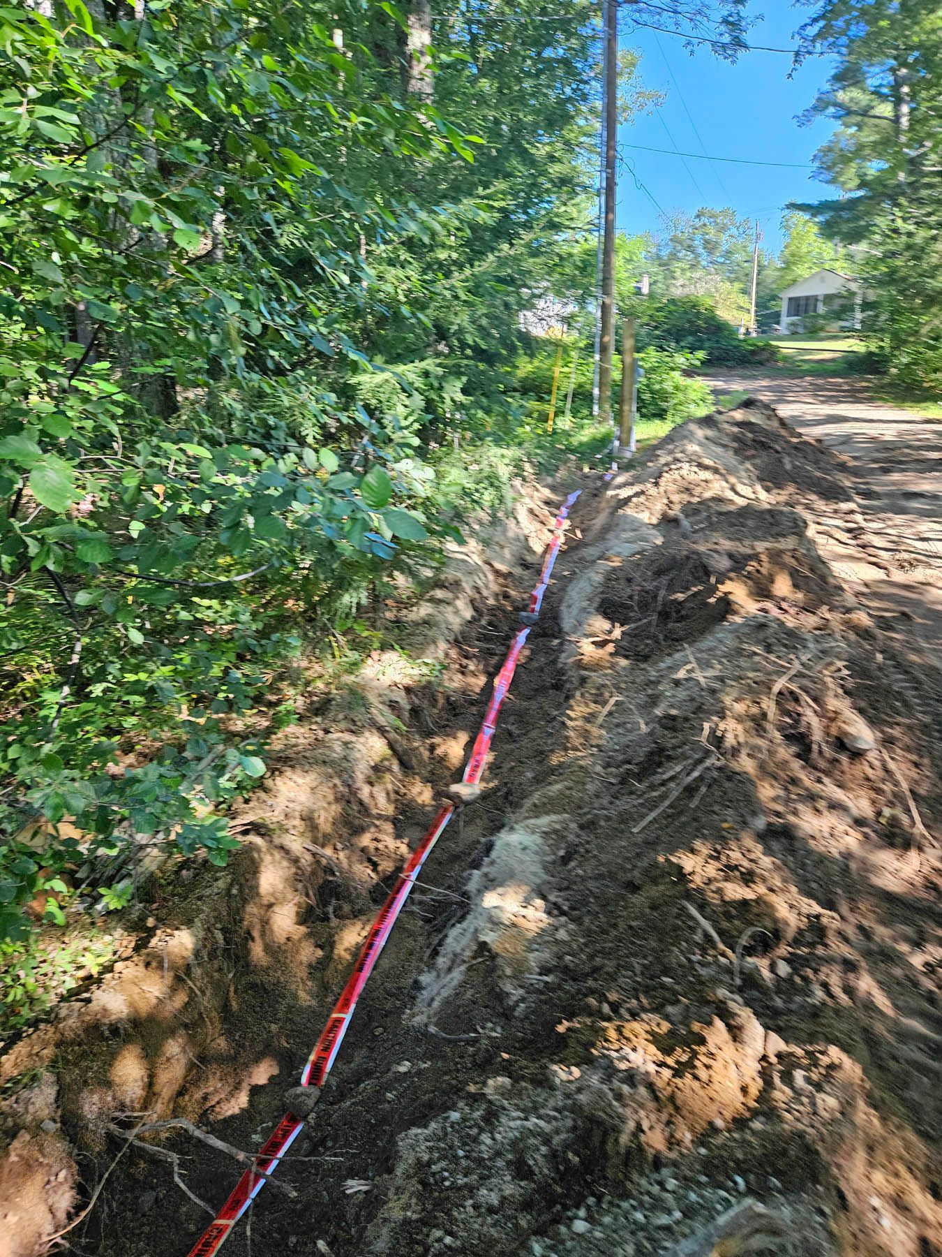 A trench with red marking tape, likely for underground utilities, beside a dirt road. Trees line the left.