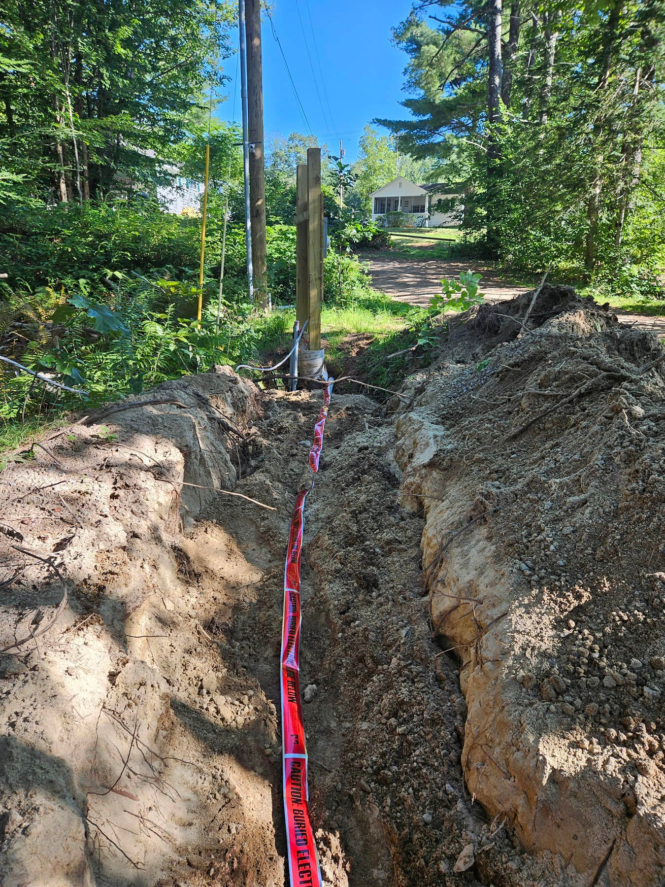 A deep trench with red caution tape; a wooden post stands in the distance near trees and a utility pole.