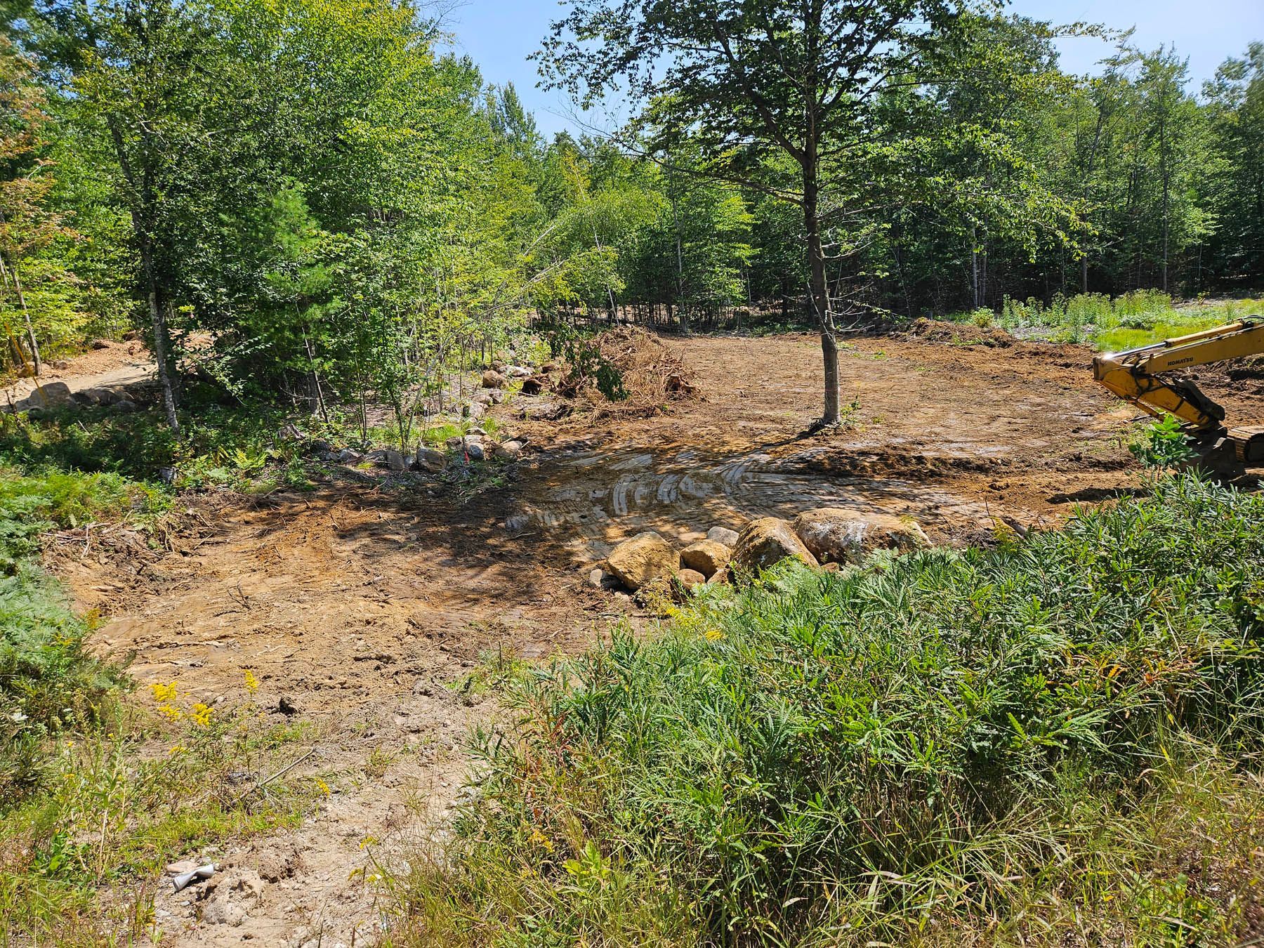 Dirt clearing in a wooded area, likely for construction. The soil is brown, with green vegetation on the edges.