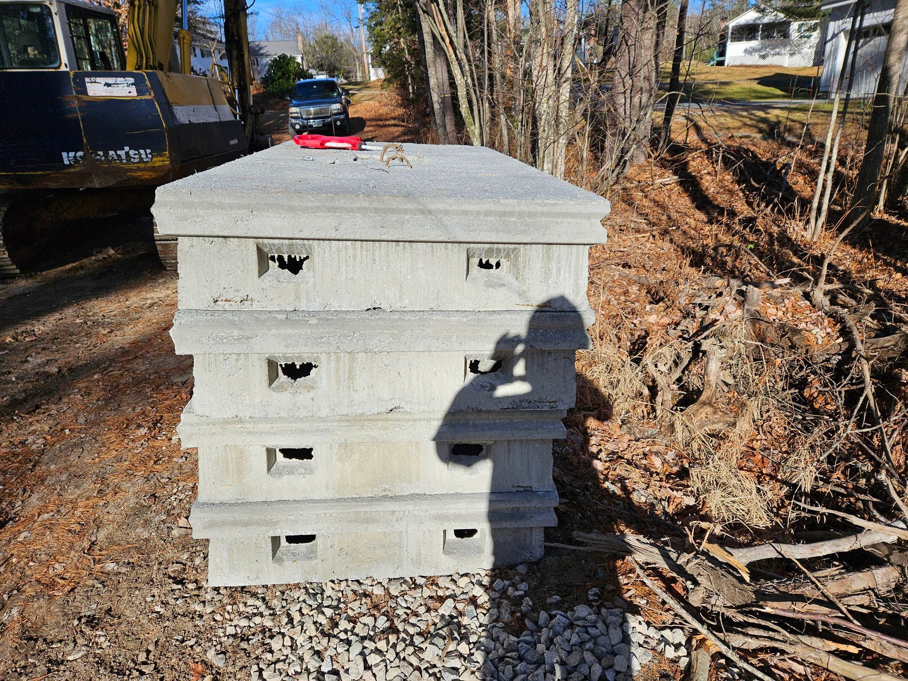 Stack of concrete utility boxes beside a gravel path, with excavator and foliage in the background.