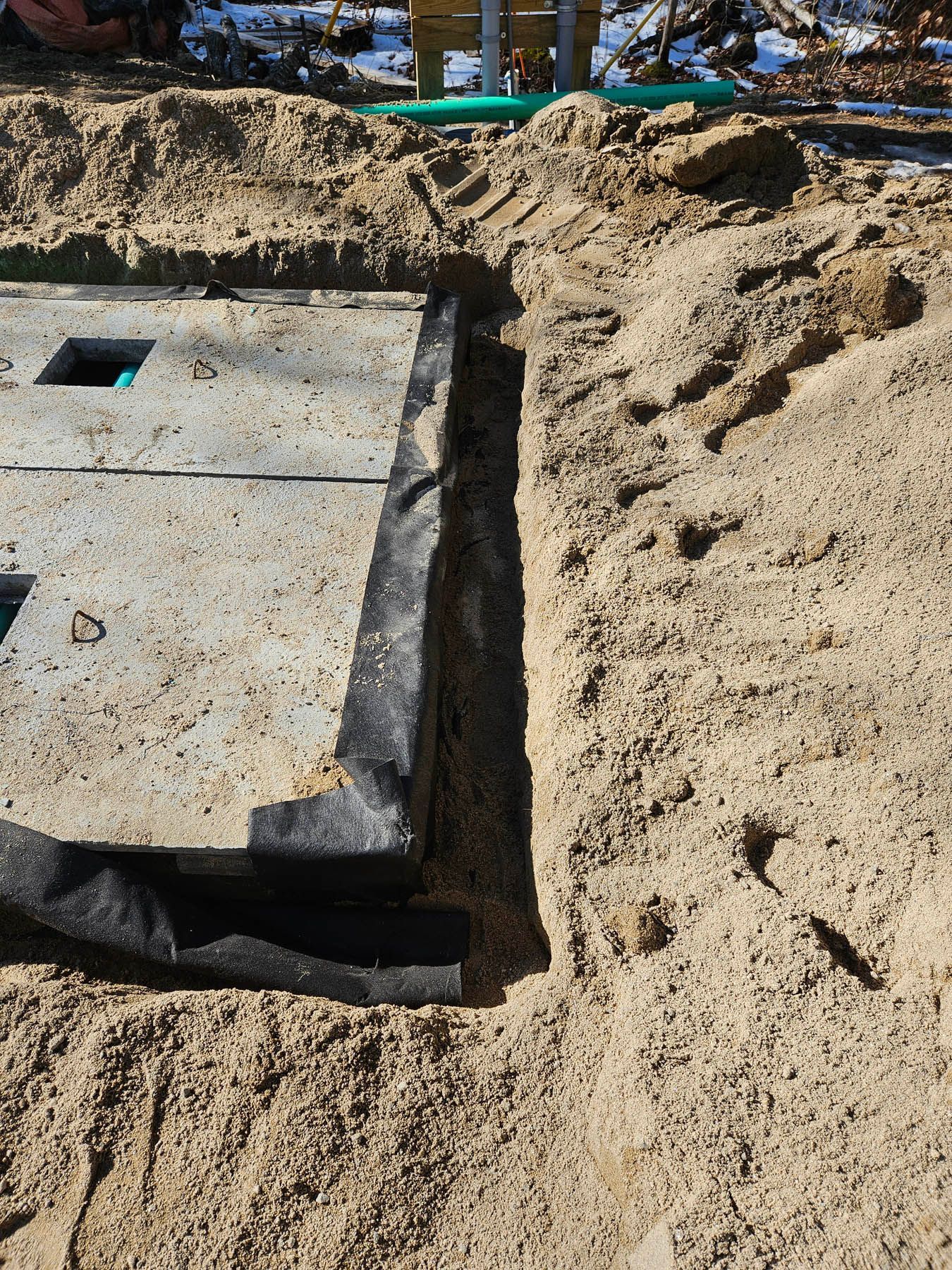 Excavated trench with black fabric lining, next to a concrete structure in a sandy environment.