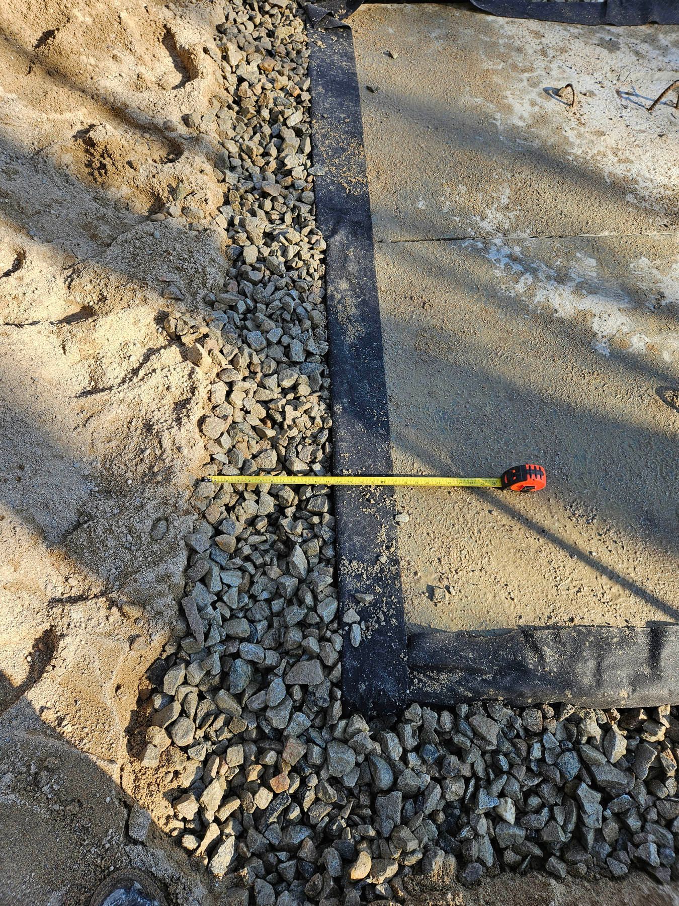Gravel border alongside concrete and sand, with a yellow tape measure extending into the gravel.
