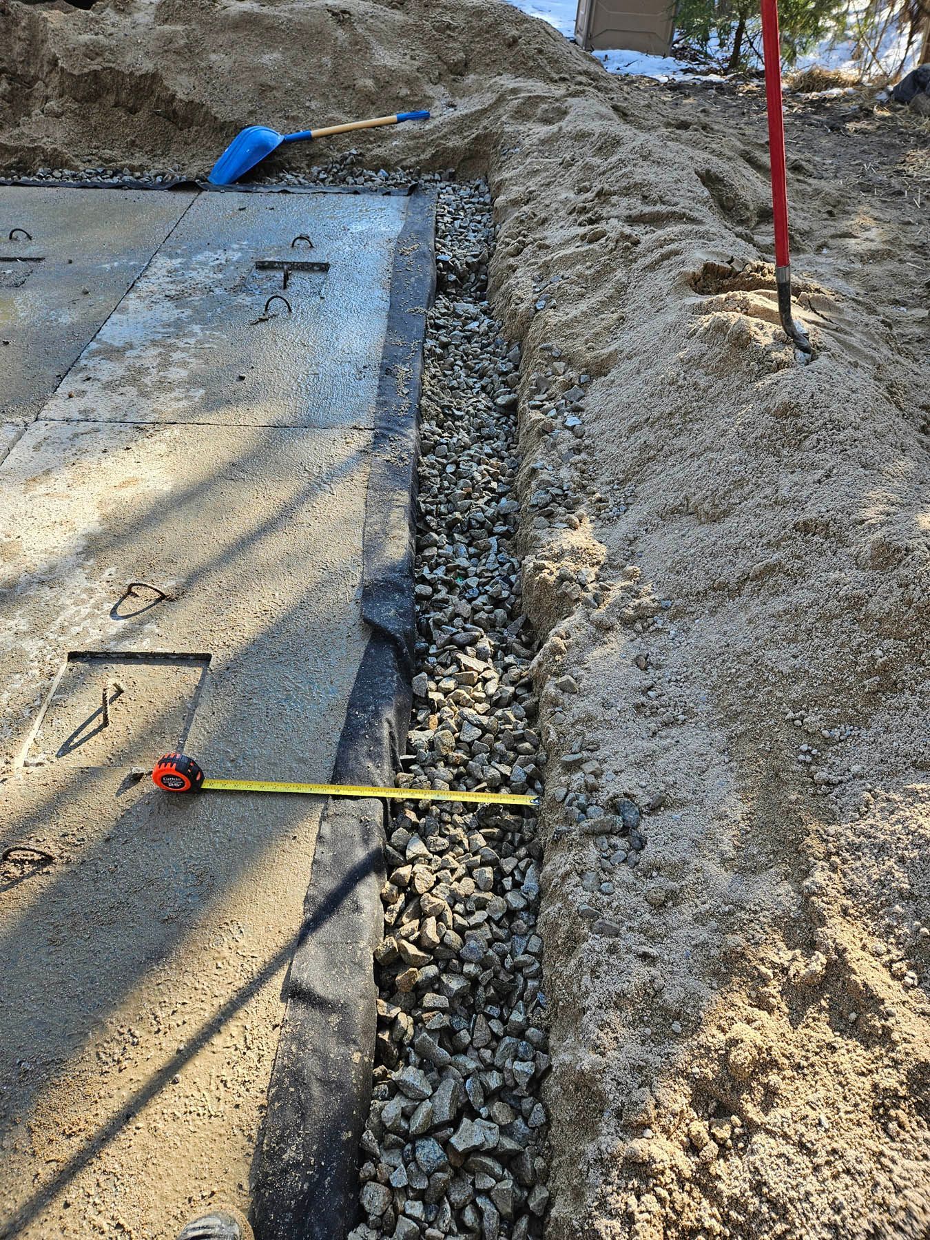 Gravel-filled trench next to a concrete slab, with a tape measure showing width, for drainage. Construction site.