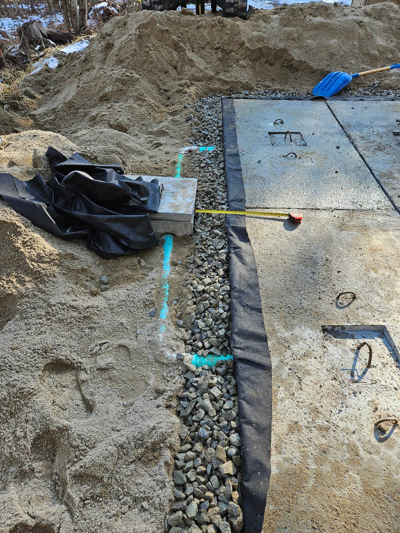 Construction site with gravel, sand, concrete forms, and black fabric edging. A blue brush lies on the surface.