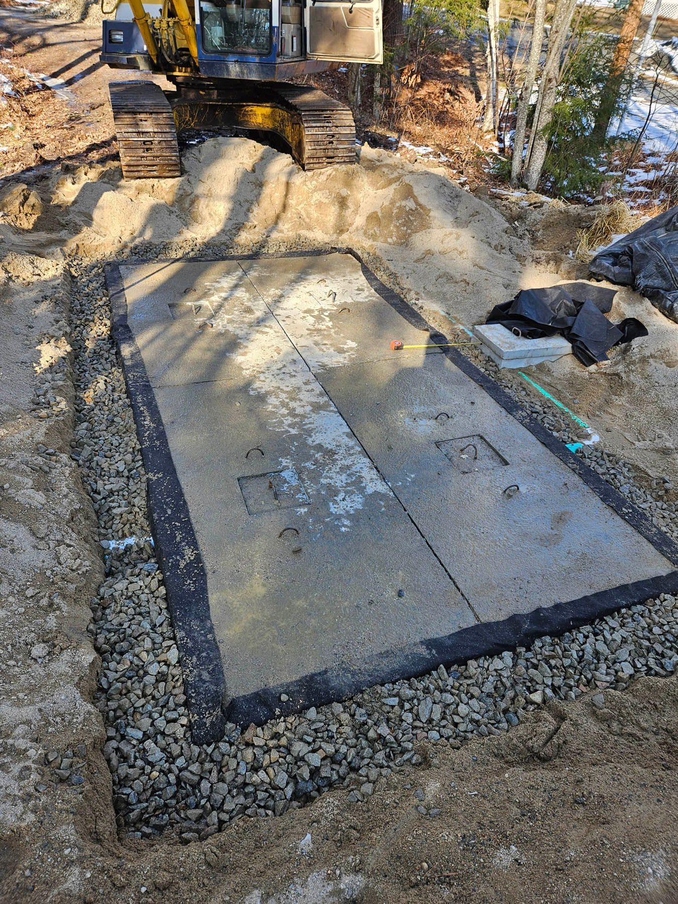 Excavator next to a concrete pad with gravel border and two access covers, outdoors.