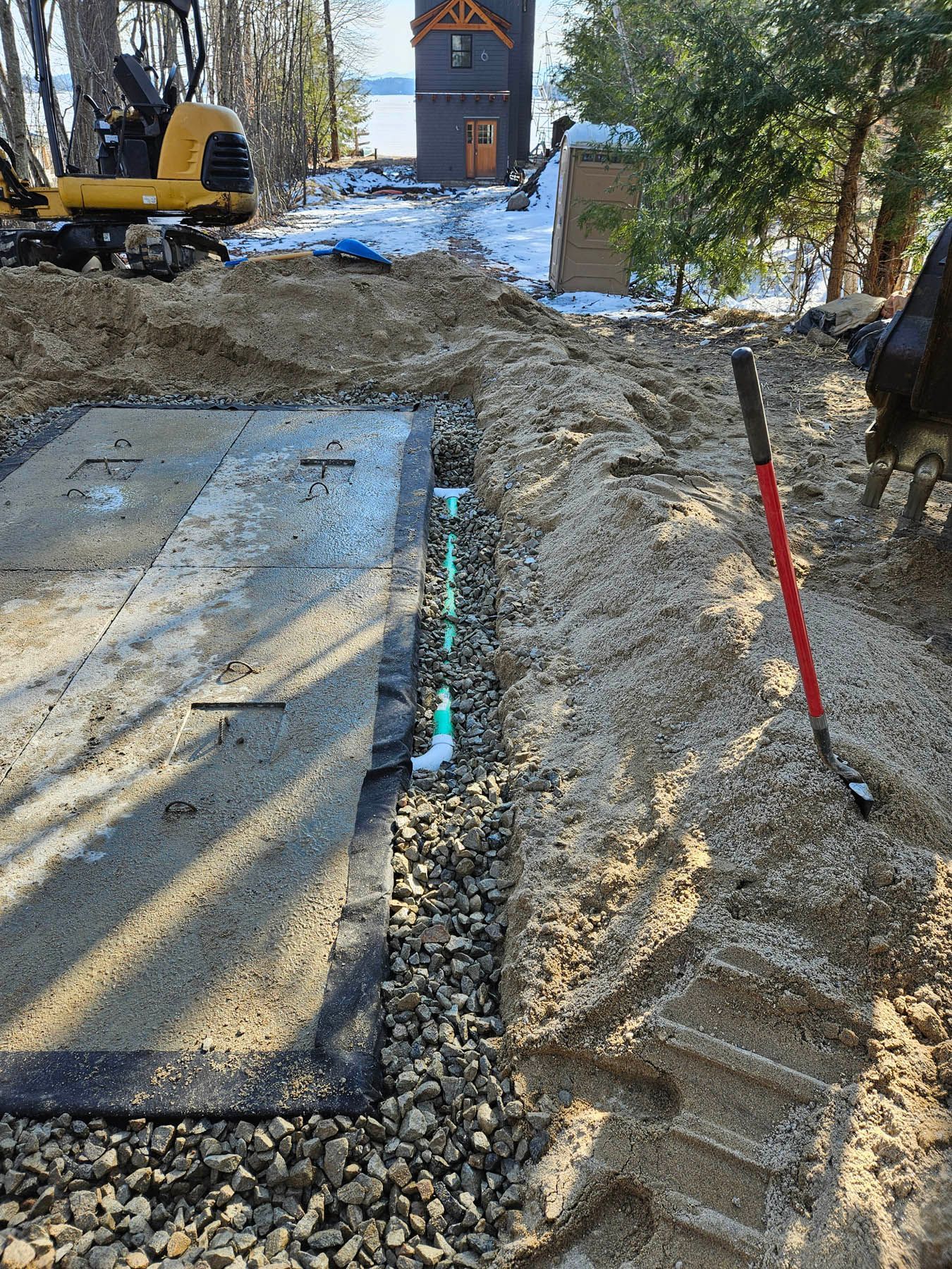 Construction site with exposed septic system components and piping, near a house and lake.