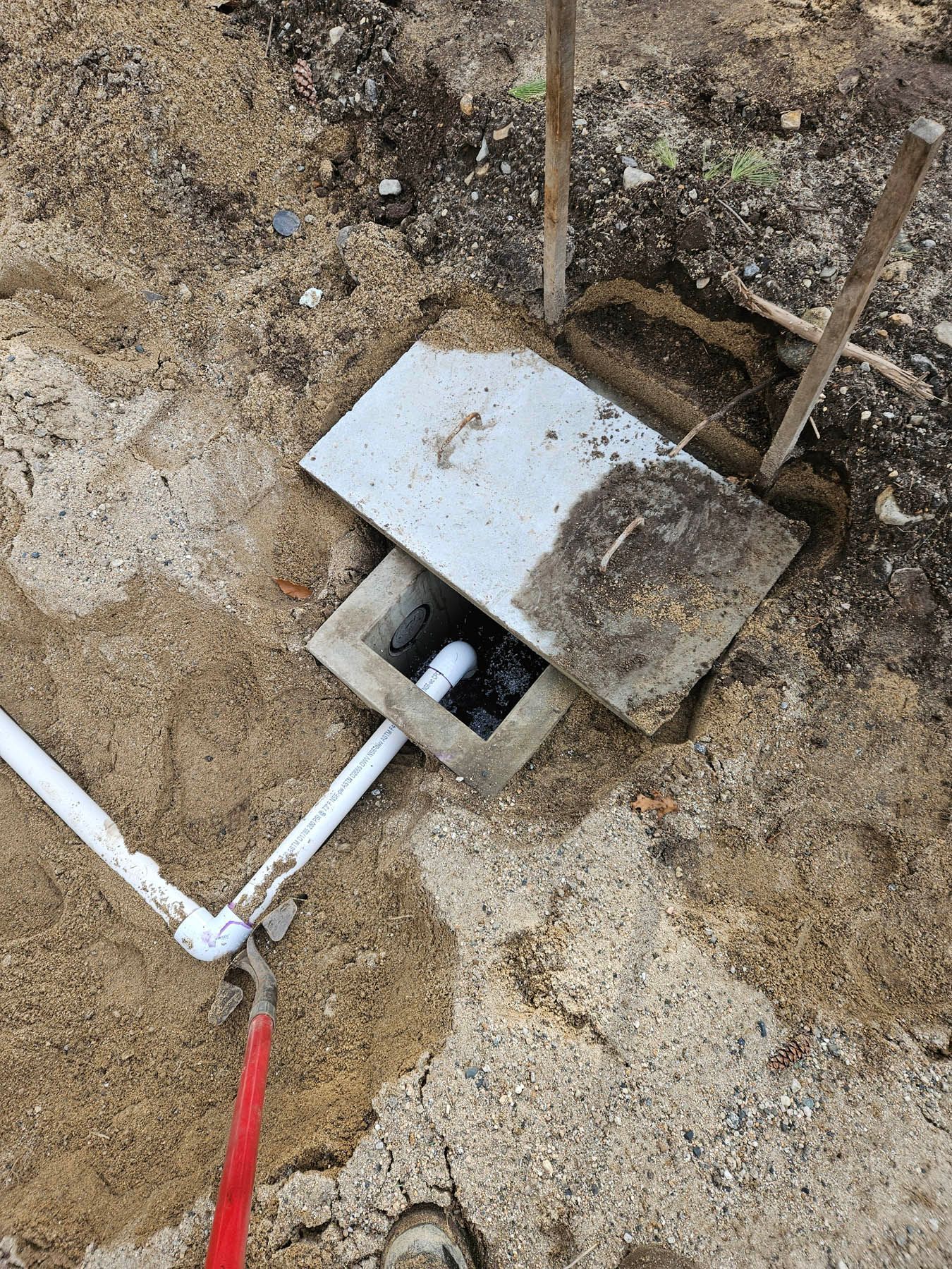 Open concrete box with white pipe connections, covered by a concrete lid, surrounded by tan dirt.