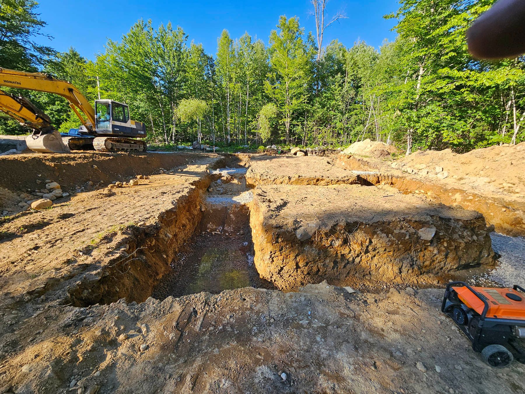 Excavation site, brown dirt, backhoe, trenches, generator, trees in background, sunny day.